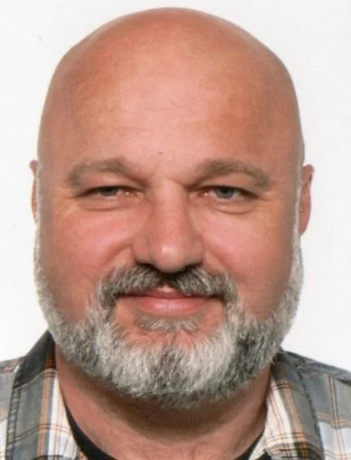 Close-up portrait of a middle-aged bald man with a gray beard, wearing a plaid shirt, smiling at the camera.