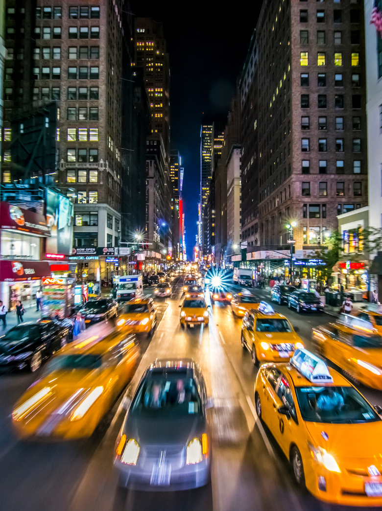 Nighttime city street filled with yellow taxis and cars, tall buildings with lit windows lining the street, downtown glow with bright city lights and skyscrapers in the background.