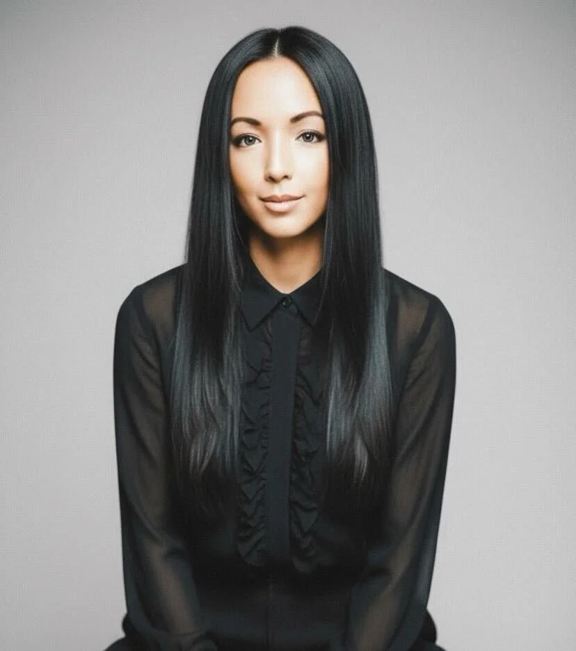 Portrait of a woman with long black hair wearing a sheer black blouse with ruffles, sitting against a plain gray background.