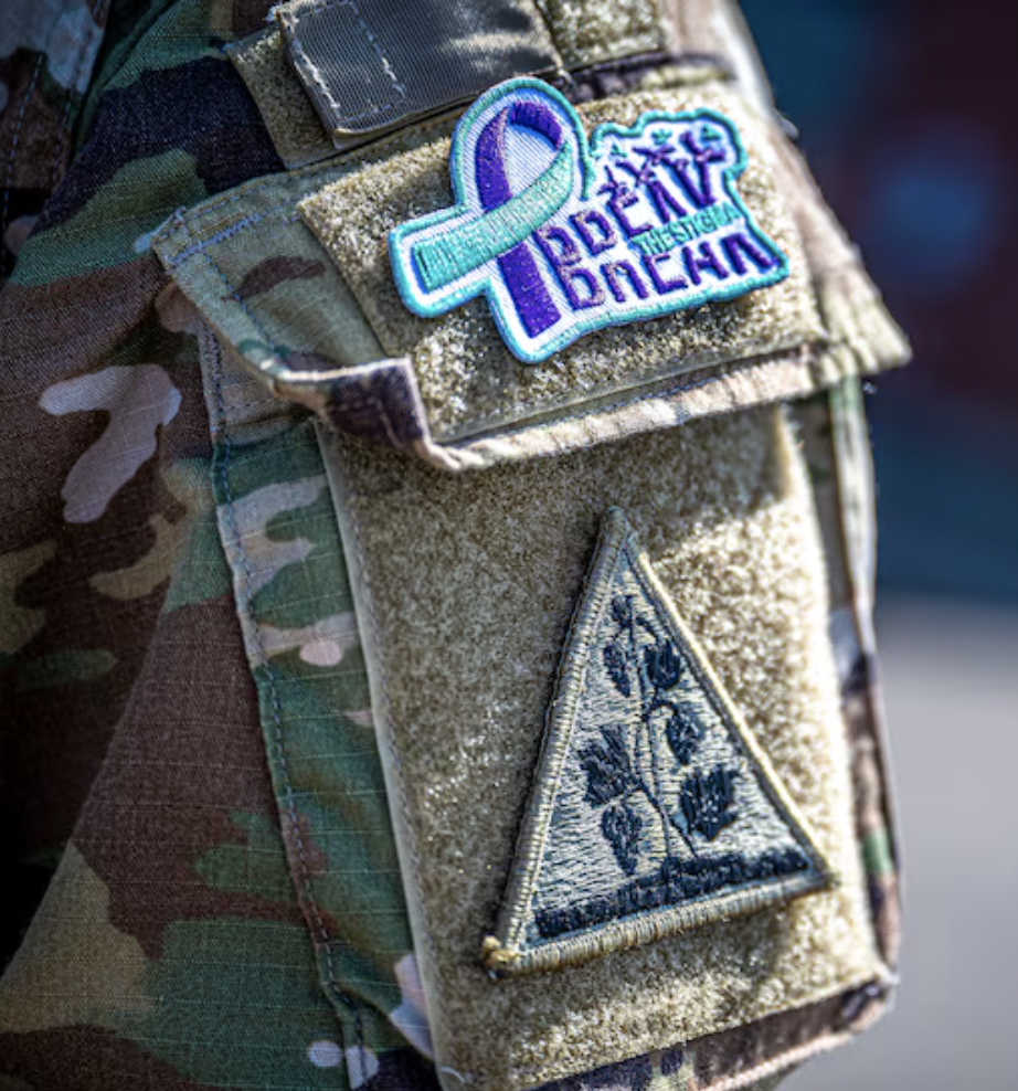 Close-up of a military uniform with patches, including a teal ribbon embroidered with the text "Break The Stigma" and a triangular patch with a laurel and crown design.