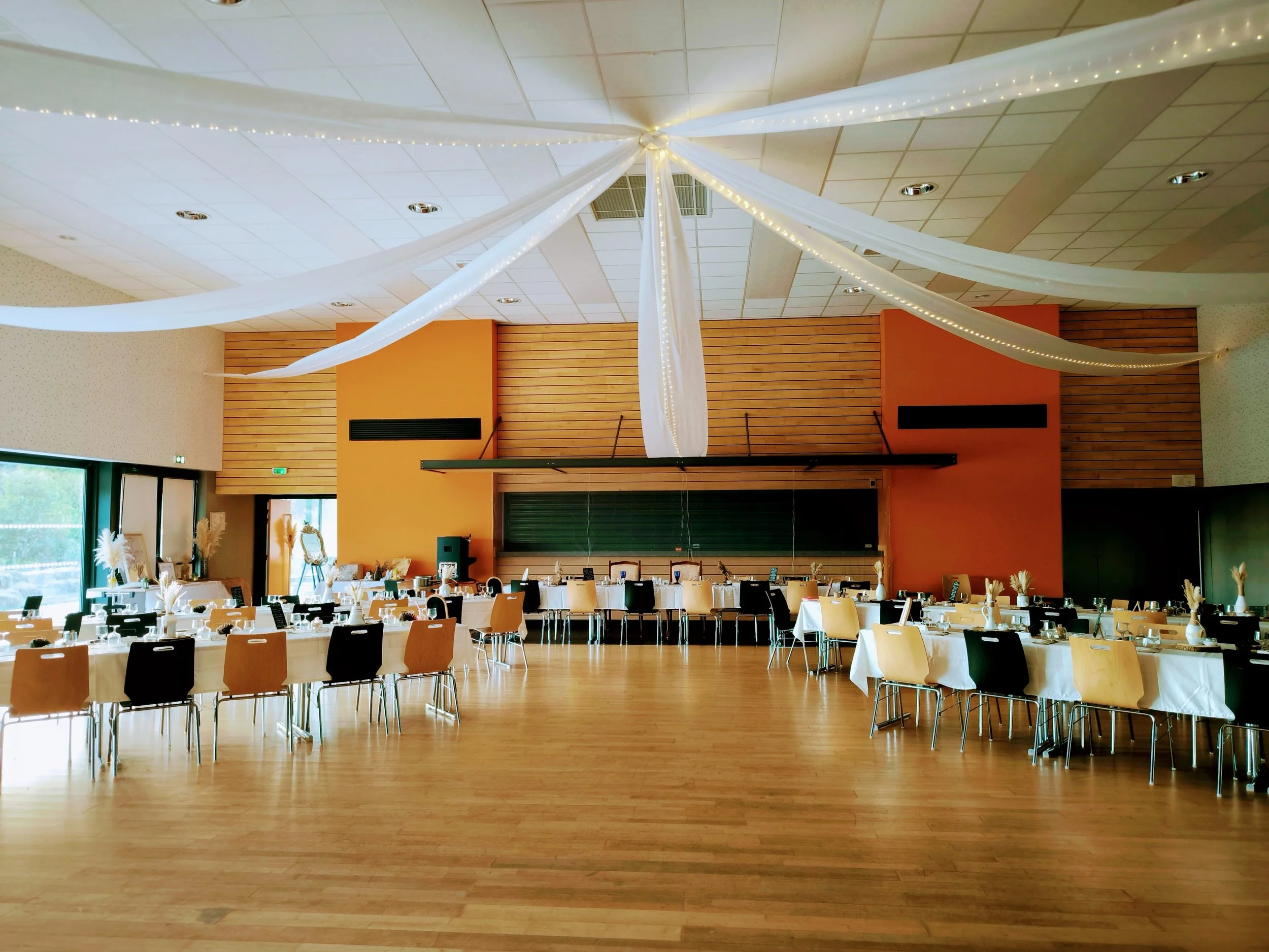 Salle de réception décorée avec des tissus blancs suspendus au plafond, tables rondes avec nappes blanches, assiettes, verres et nappes, décorations florales.