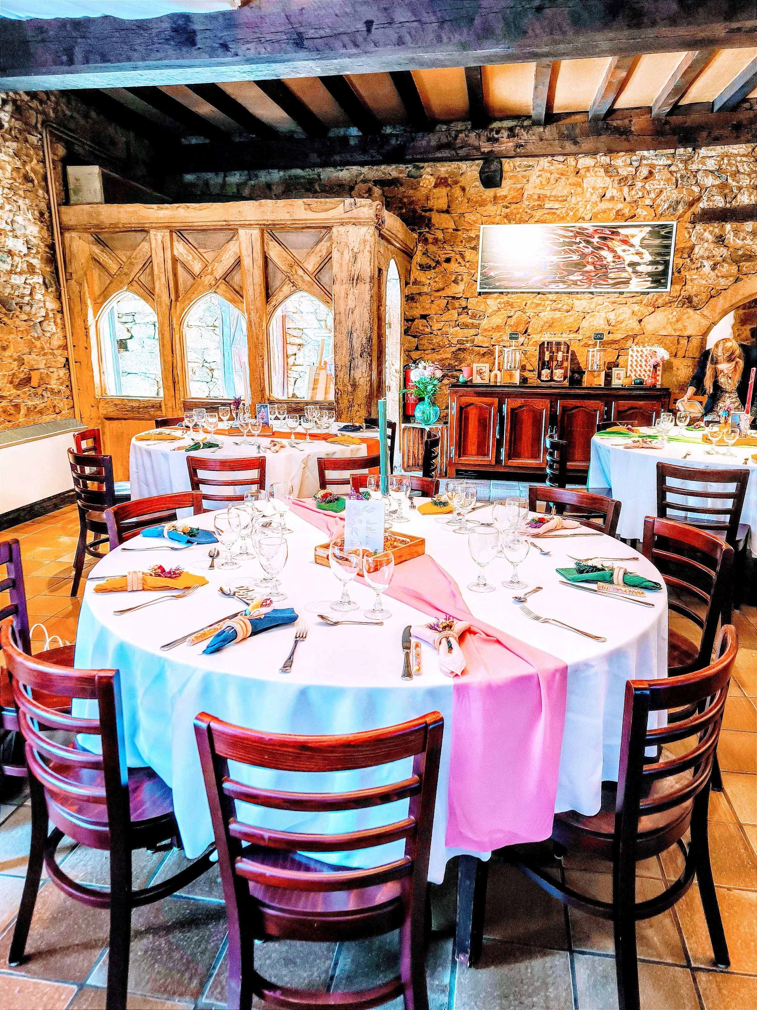 Salle de réception décorée pour un événement, avec une table ronde dressée avec des verres, assiettes, serviettes colorées et une décoration centrale, mur en pierre et mobilier en bois