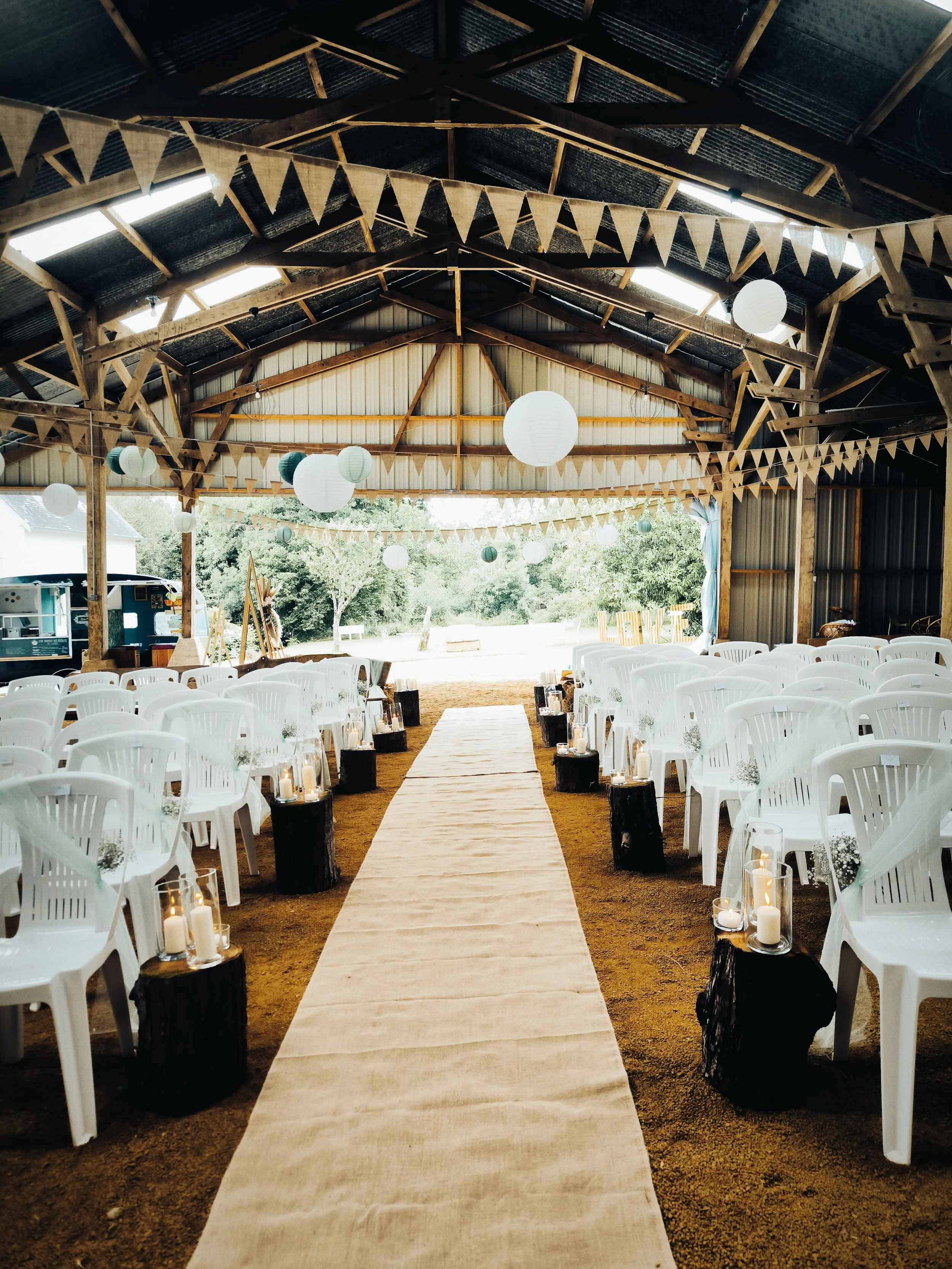 Décoration de mariage en salle en plein air avec un tapis beige au centre, des lanternes avec bougies sur des troncs d'arbres noirs, des chaises blanches, des lanternes suspendues, et des guirlandes de fanions en tissu.