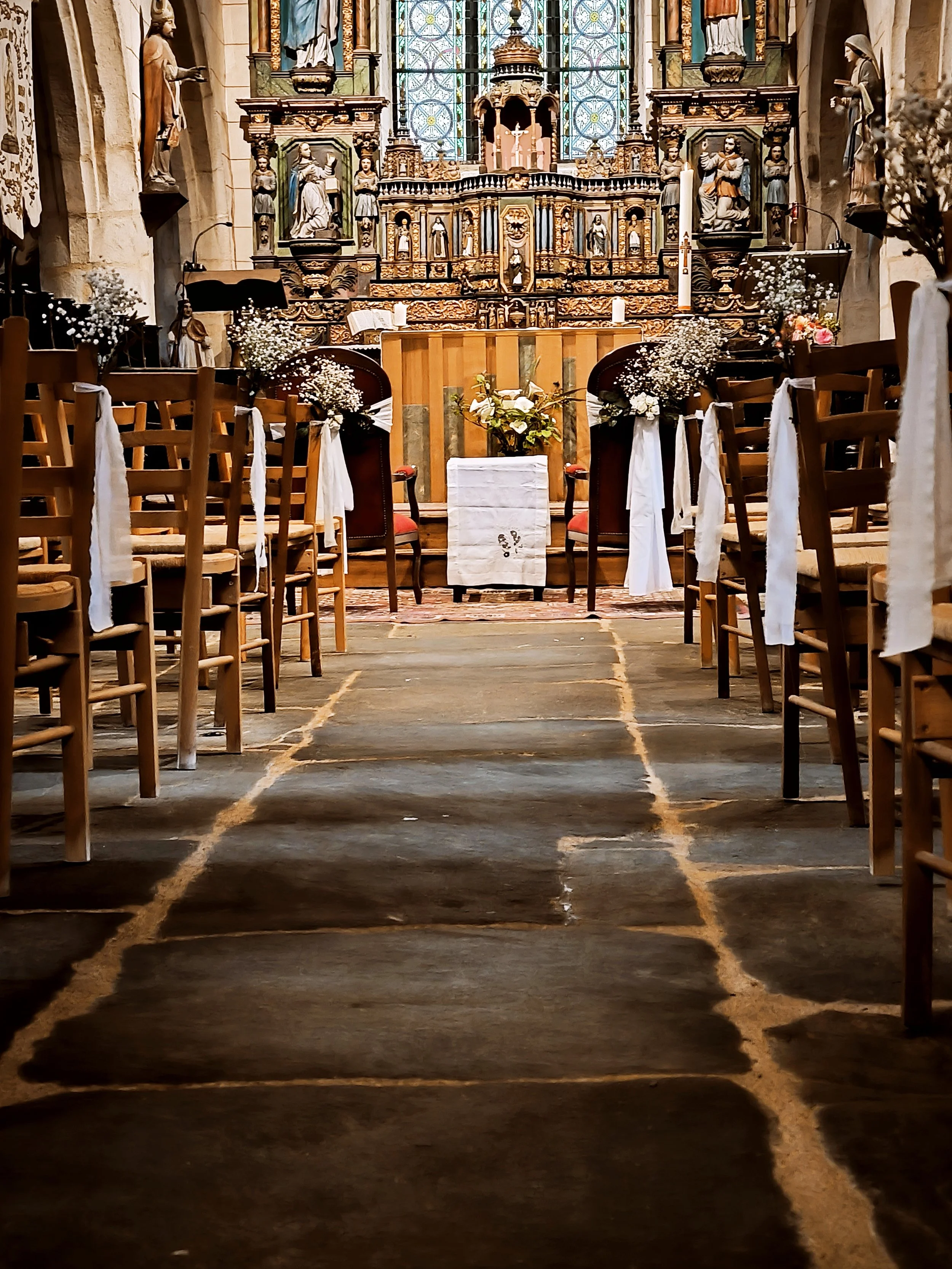 Intérieur d'une église ornée, toilet créée pour un mariage avec des chaises et des fleurs, altar en bois avec statues et vitraux colorés en arrière-plan.