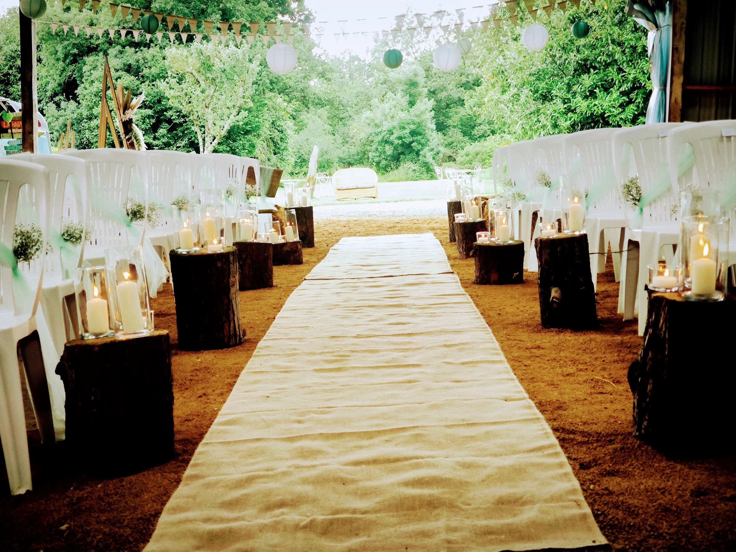 Allée de mariage en extérieur avec chaises blanches décorées de rubans, lanternes et bougies sur des troncs en bois, sous un toit de lampions, avec un fond de verdure.