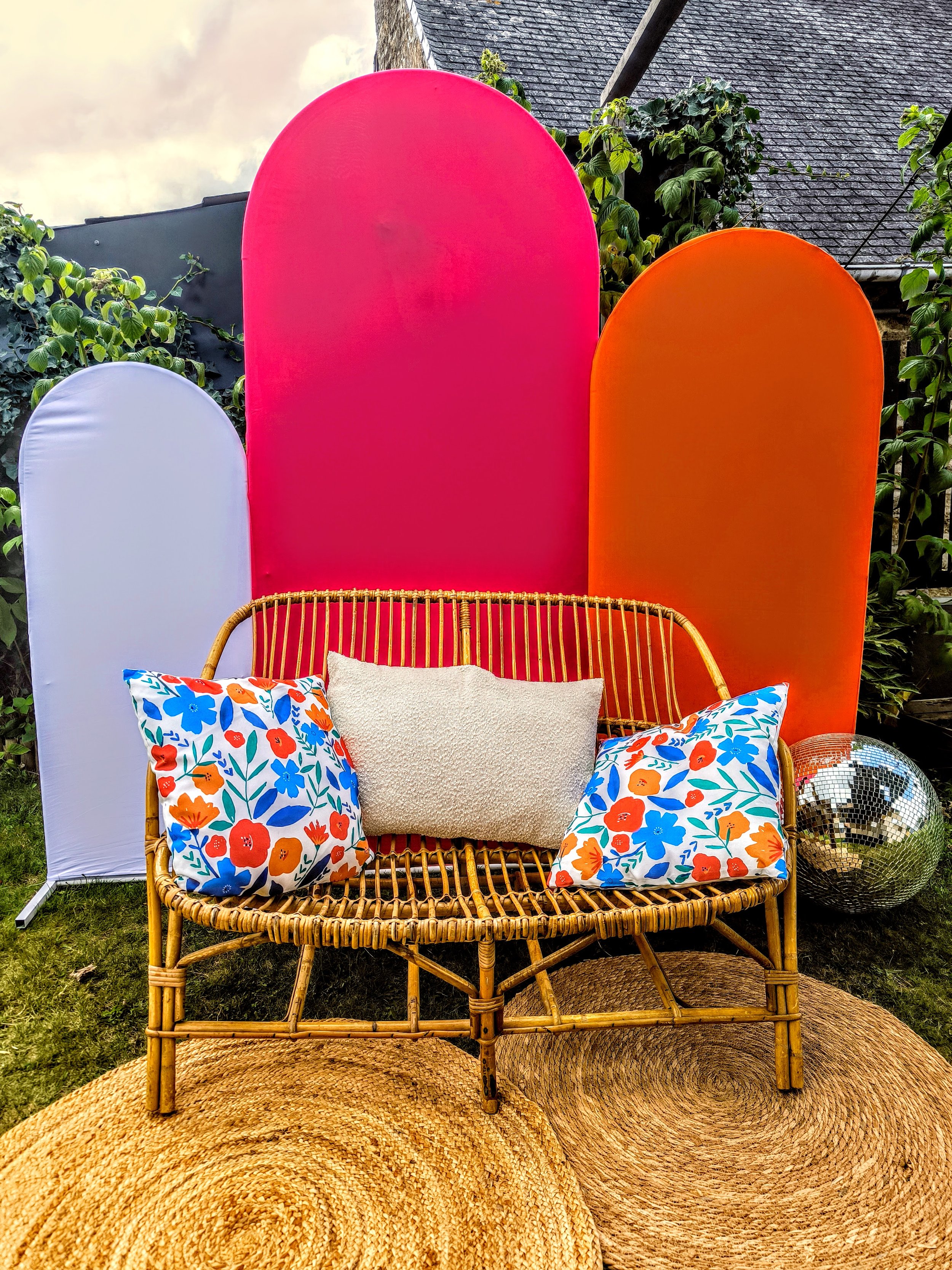 Décor extérieur avec une banquette en rotin, deux coussins aux motifs floraux colorés, un tapis en jute, une boule de disco réfléchissant, un fond de panneaux colorés rose fuchsia, orange, blanc.