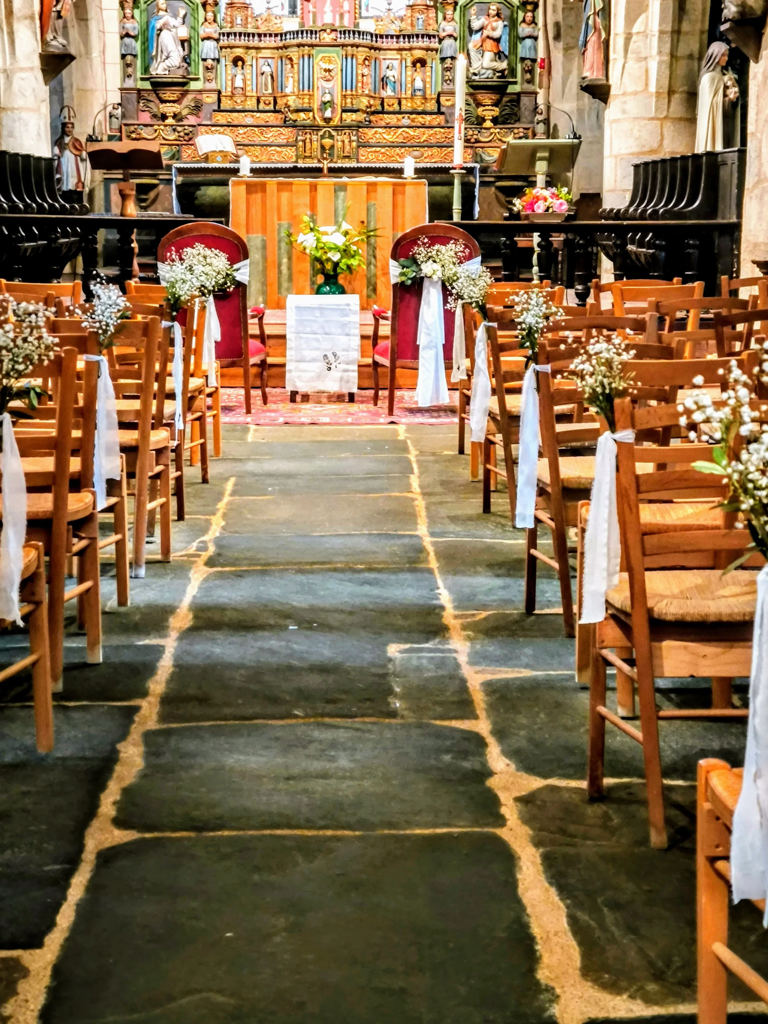 Intérieur d'une église décorée pour un mariage, des chaises en bois avec des rubans blancs et des fleurs fraiches, et un autel orné de fleurs.