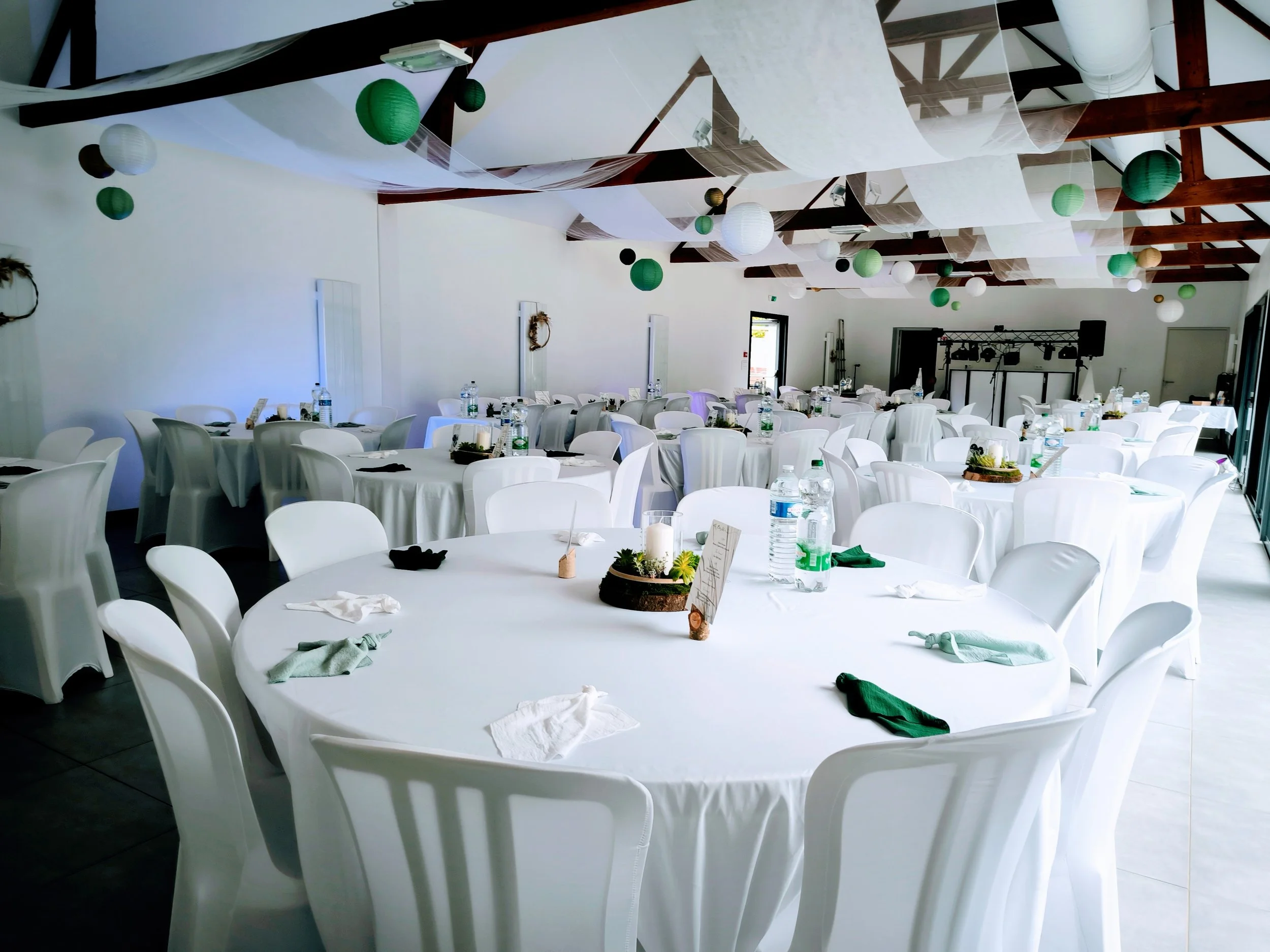 Salle de réception décorée avec des tables rondes et des chaises blanches, lampions verts et blancs suspendus au plafond, tables avec bouteilles d'eau, serviettes, et petits centres de table.