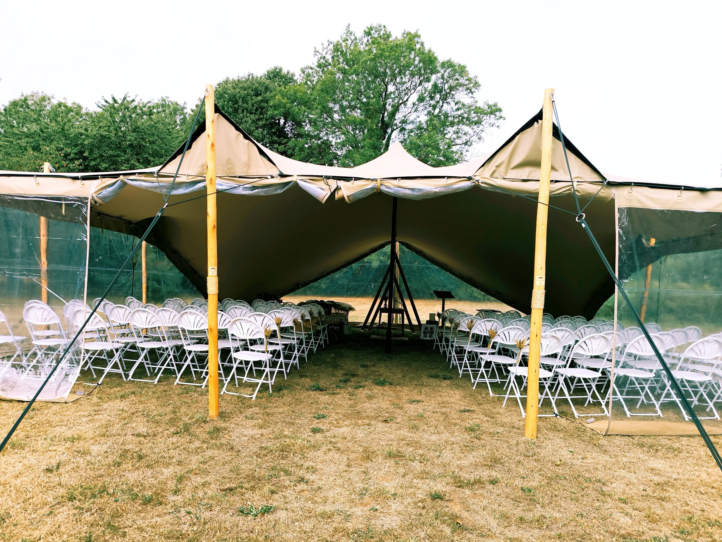 Une tente blanche installée en extérieur avec des chaises blanches en rangée à l'intérieur, probablement pour une cérémonie laïque, avec un arrière-plan d'arbres et de ciel clair.