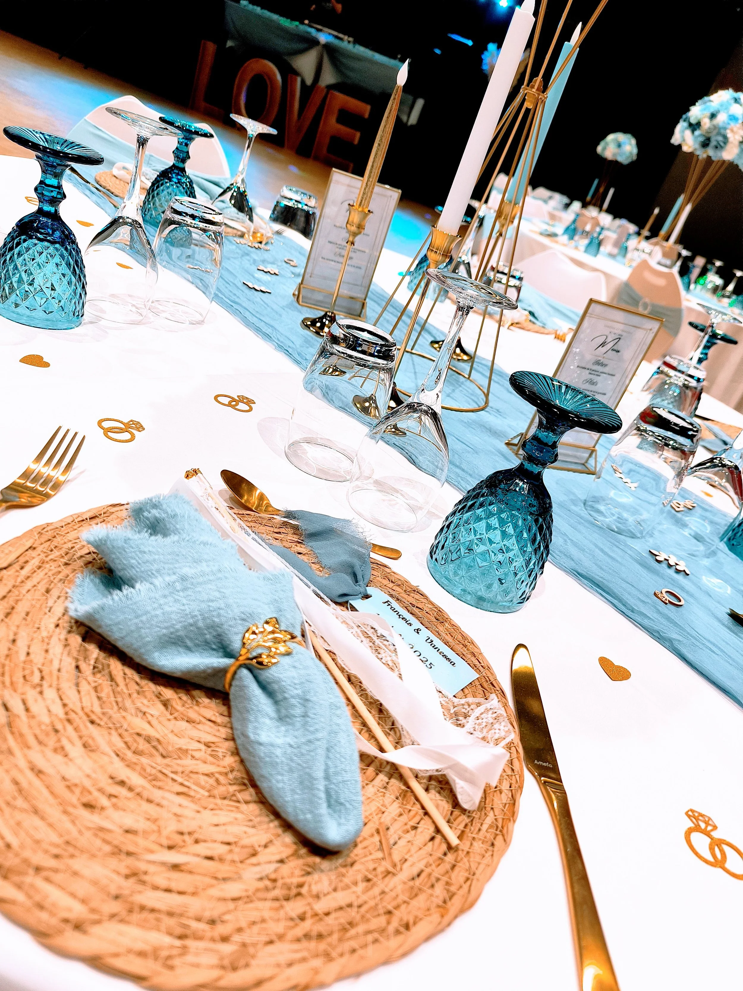 Table décorée avec des verres bleus, des bougeoirs en or avec des chandelles, une nappe blanche avec un chemin de table bleu, des assiettes en bambou, et des couverts en or, pour un mariage ou un événement spécial. Inclut aussi des petits cœurs en or