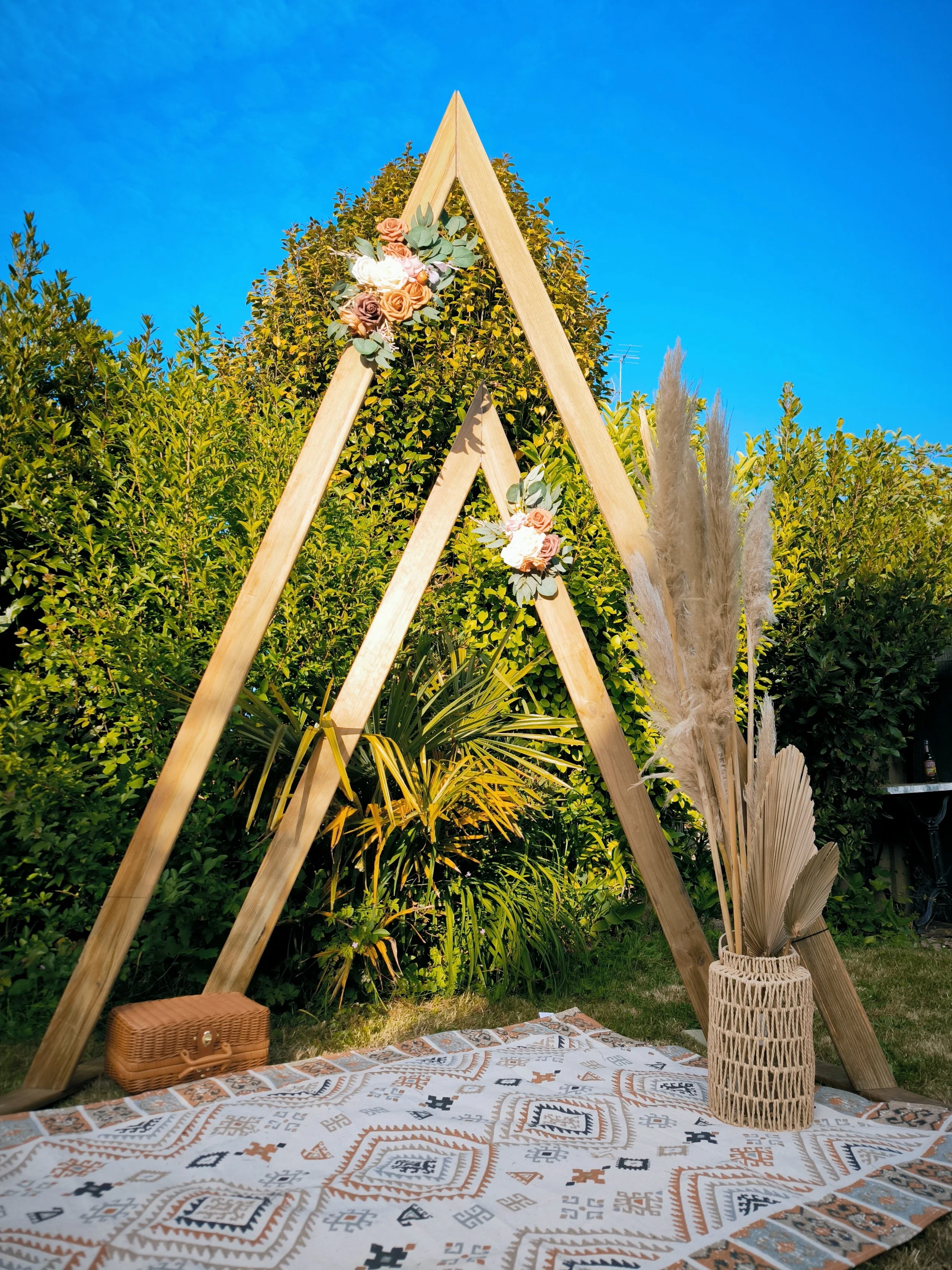 Décor de mariage en plein air avec un cadre en bois en forme de tipi, décoré de fleurs, un tapis à motifs, un panier en osier avec des plantes séchées et un petit coffret en bois, le tout sous un ciel bleu avec des buissons en arrière-plan.
