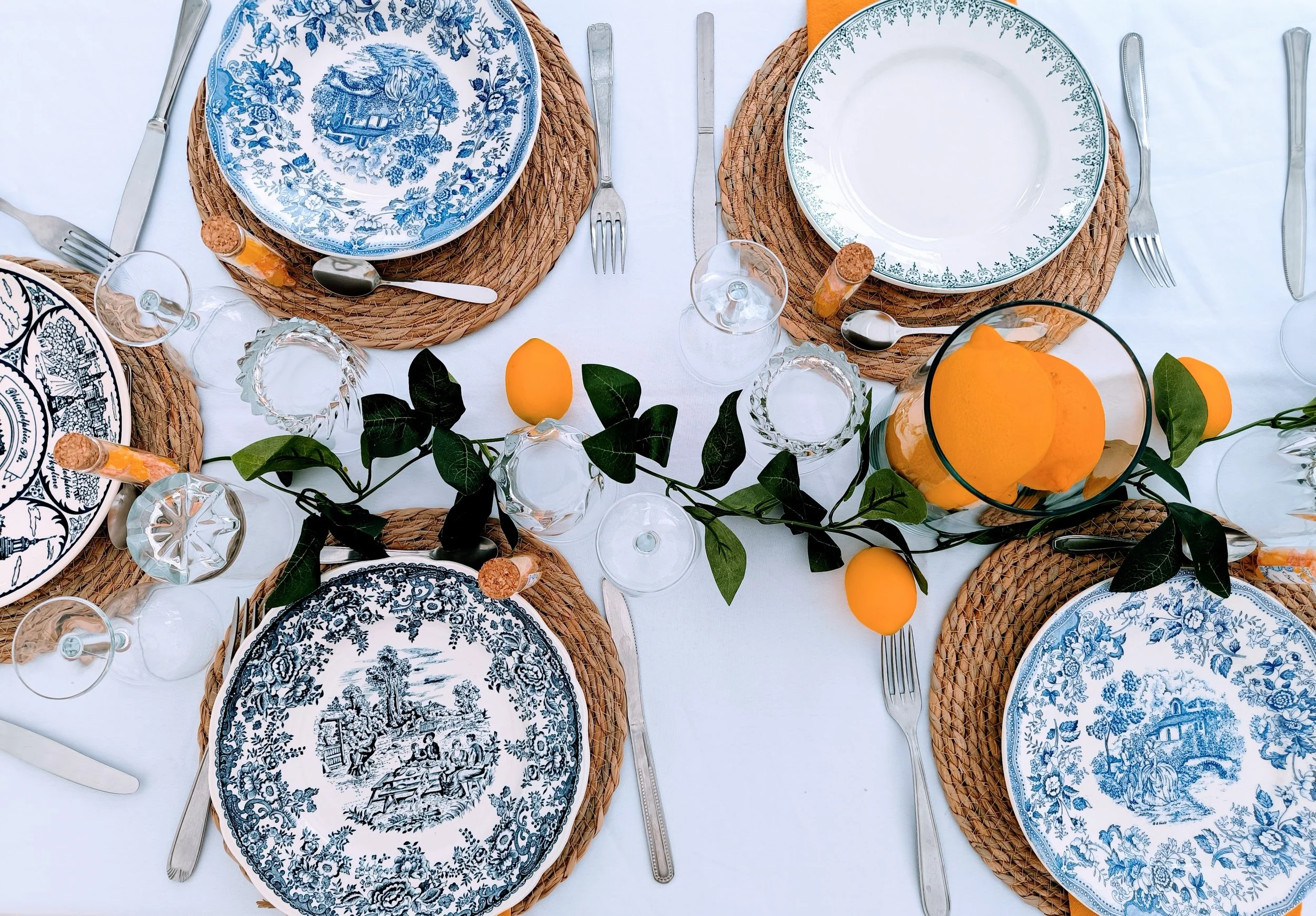 Table dressée style méditerranéen avec des assiettes en porcelaine bleu et blanc, cœurs en verre, couverts en métal, décorations de feuilles vertes, citron et orange frais dans un vase.