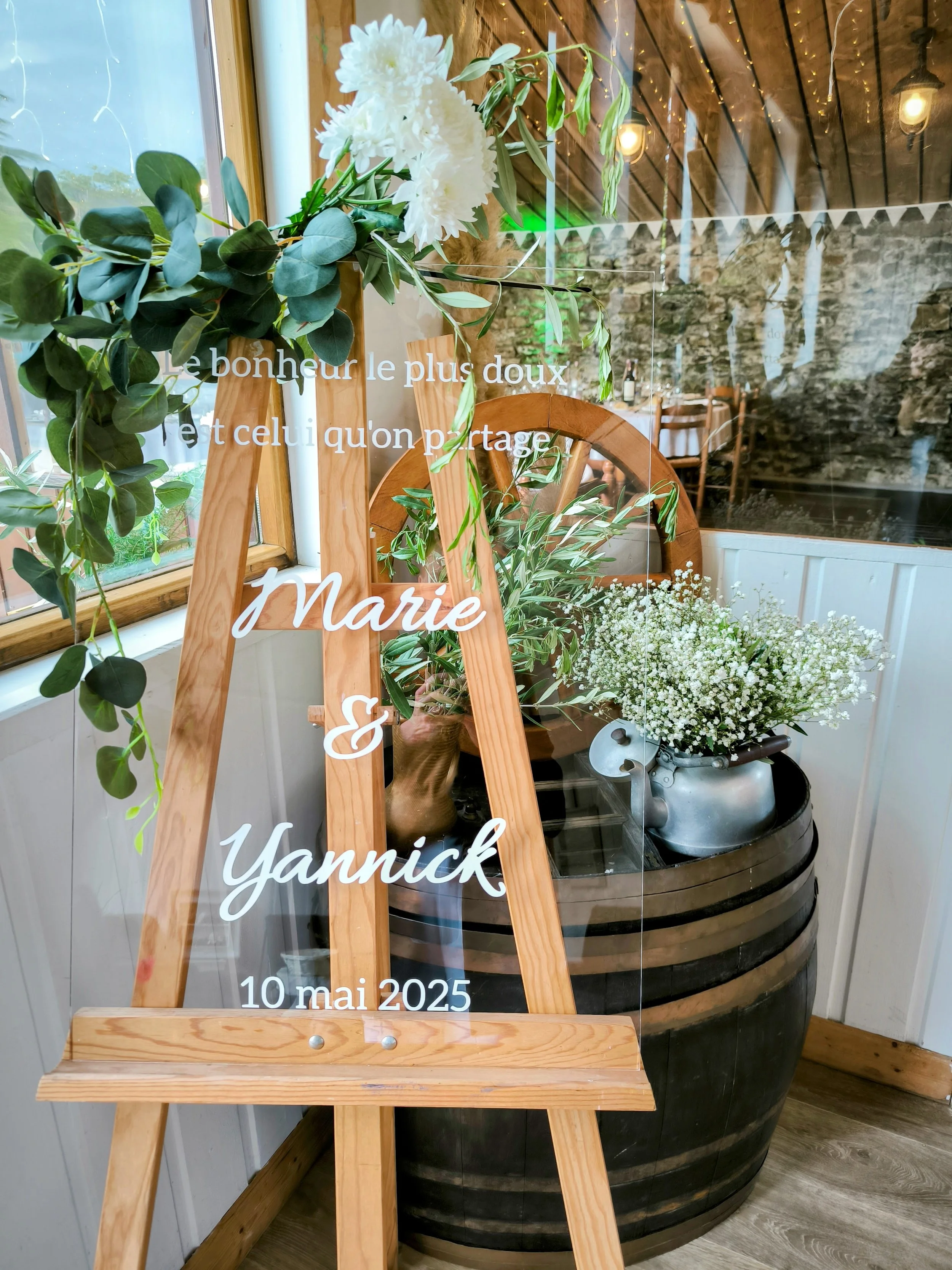 Une affiche en bois et en plexi avec des fleurs et une citation en français. La citation dit : 'Le bonheur le plus doux est celui qu'on partage', suivi des noms 'Marie & Yannick' et la date '10 mai 2025'.