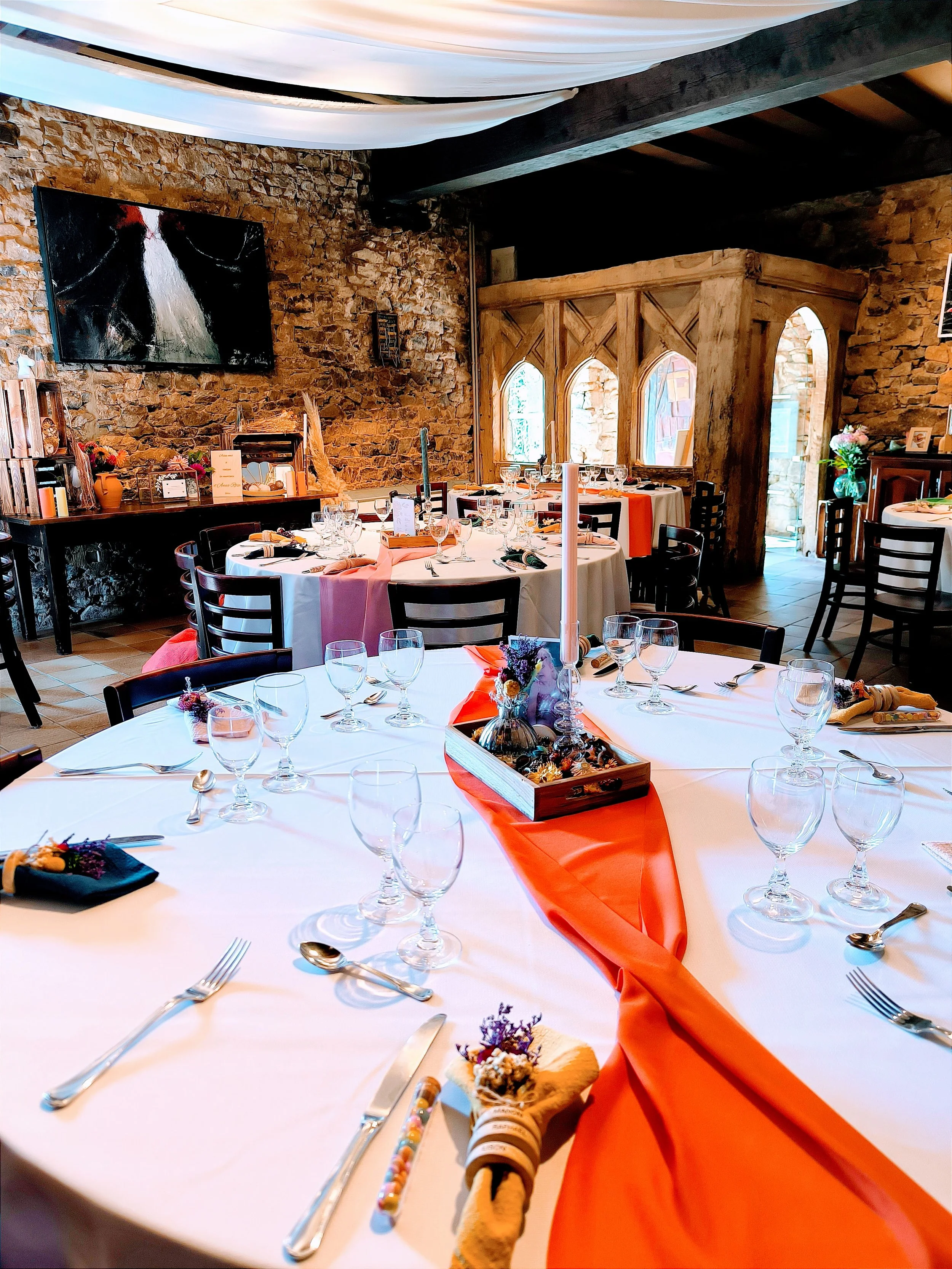 Salle de réception joliment décorée avec des tables rondes, nappes blanches, décorations florales, bougies, et un mur en pierre. Il y a aussi des fenêtres en arc et une structure en bois derrière.