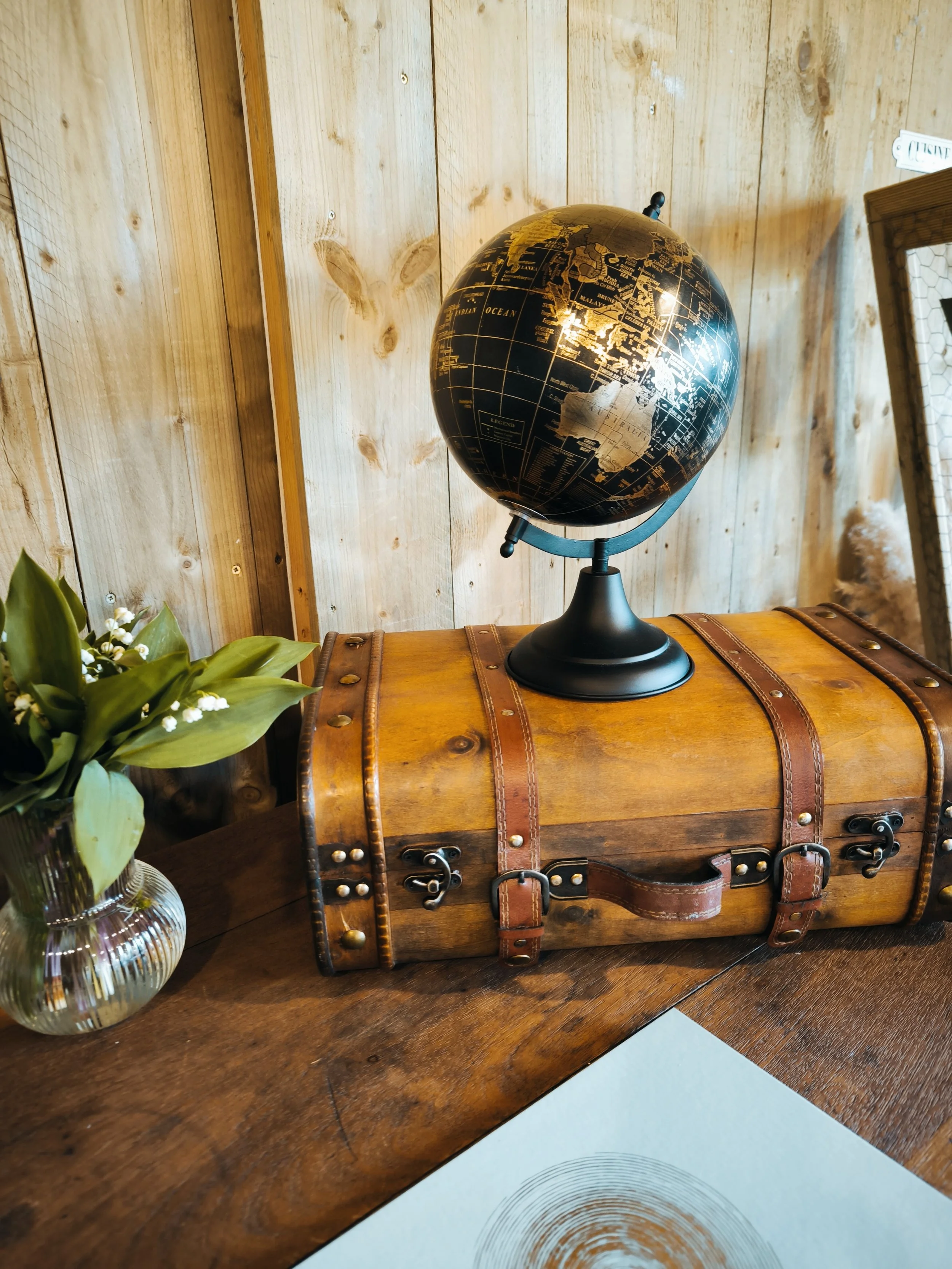 Globe terrestre noir avec des continents en or, placé sur une table en bois à côté d'une petite valise en cuir et d'un vase avec des fleurs vertes.