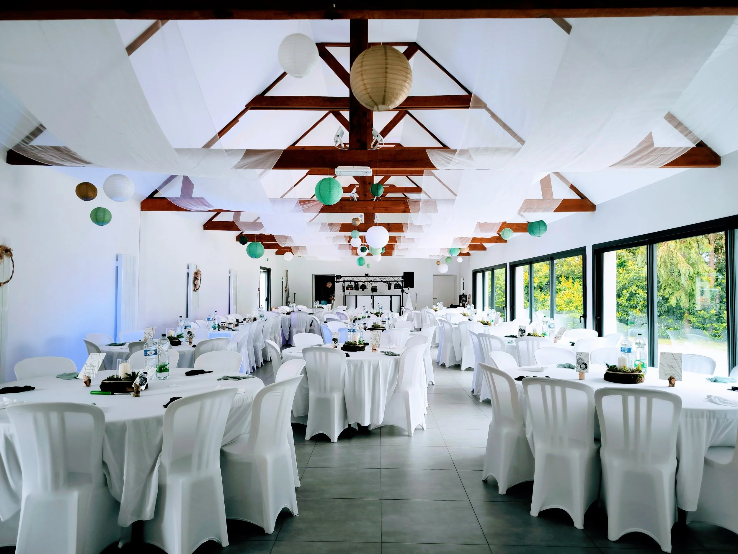 Salle de réception décorée pour un événement avec tables rondes drapées de blanc, chaises blanches, lanternes en papier suspendues au plafond, grande baie vitrée laissant entrer de la lumière naturelle, et un espace au fond avec du matériel de son et