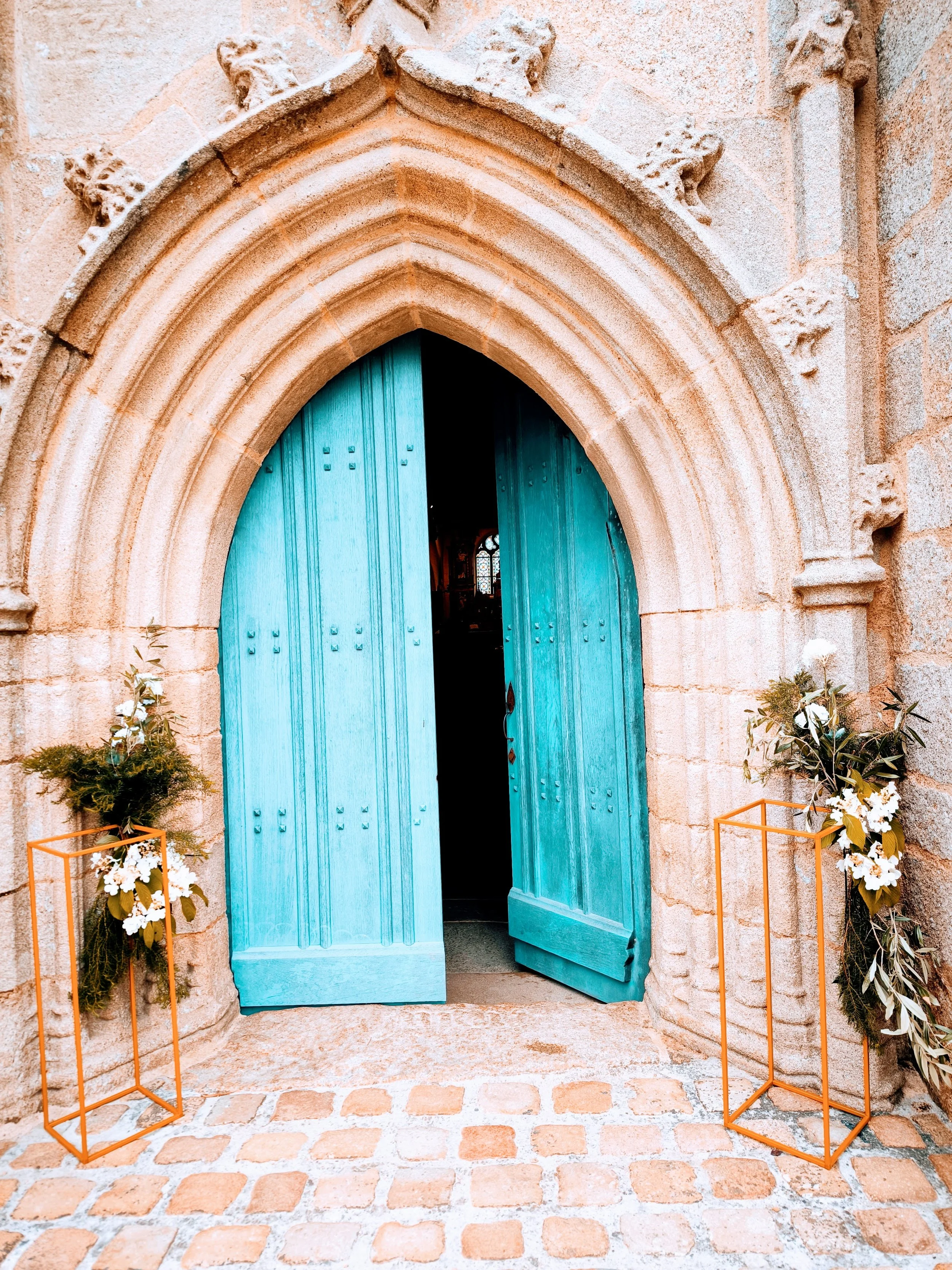 Une porte en bois turquoise entrouverte dans une entrée d'église en pierre, décorée avec des fleurs dans des supports or.