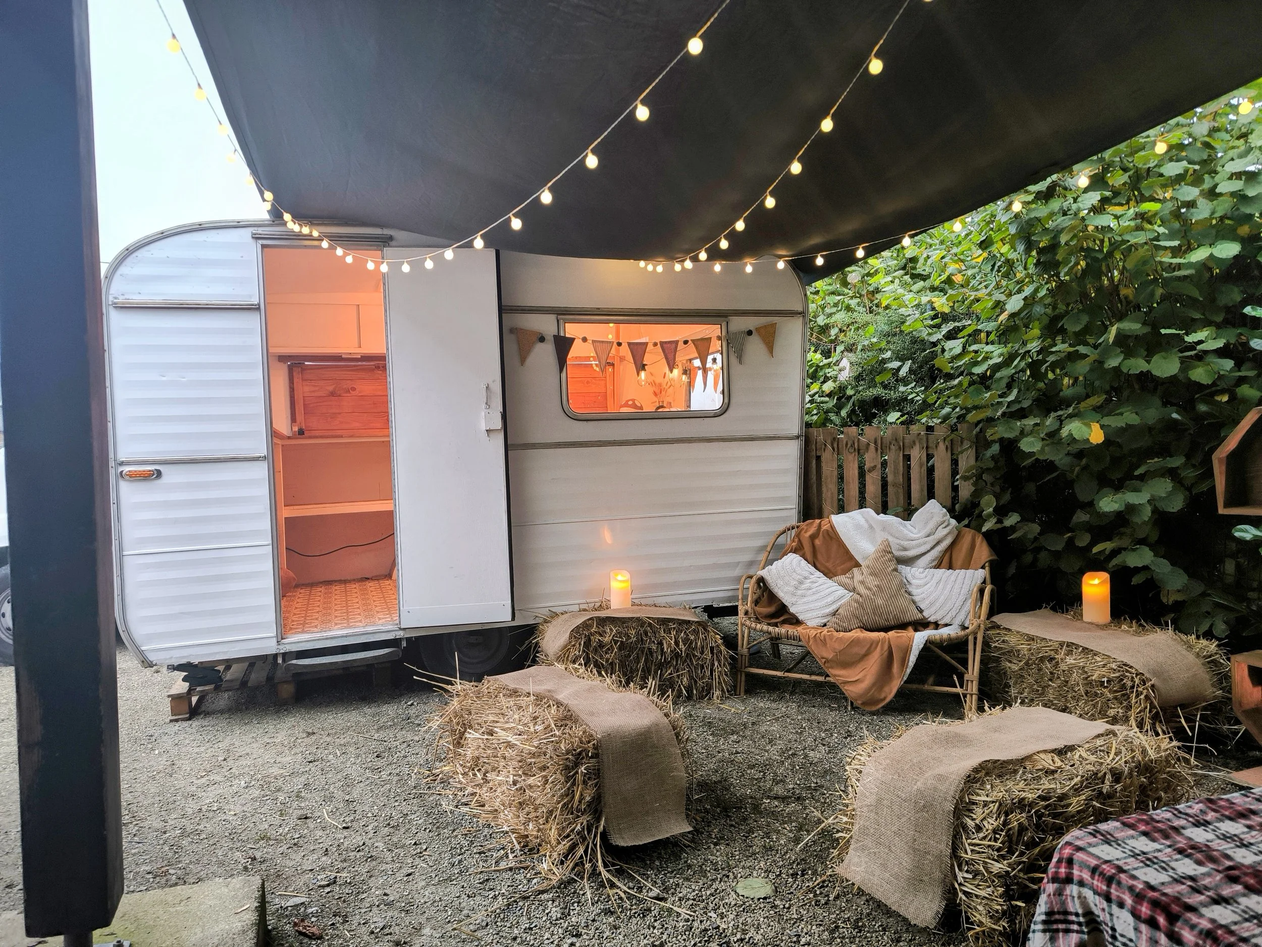 Une scène extérieure avec une caravane aménagé en photobooth, décoré de guirlandes lumineuses et des bougies. Un canapé en osier avec des coussins et des couvertures est placé devant le van, entouré de balles de foin avec des tissus. 