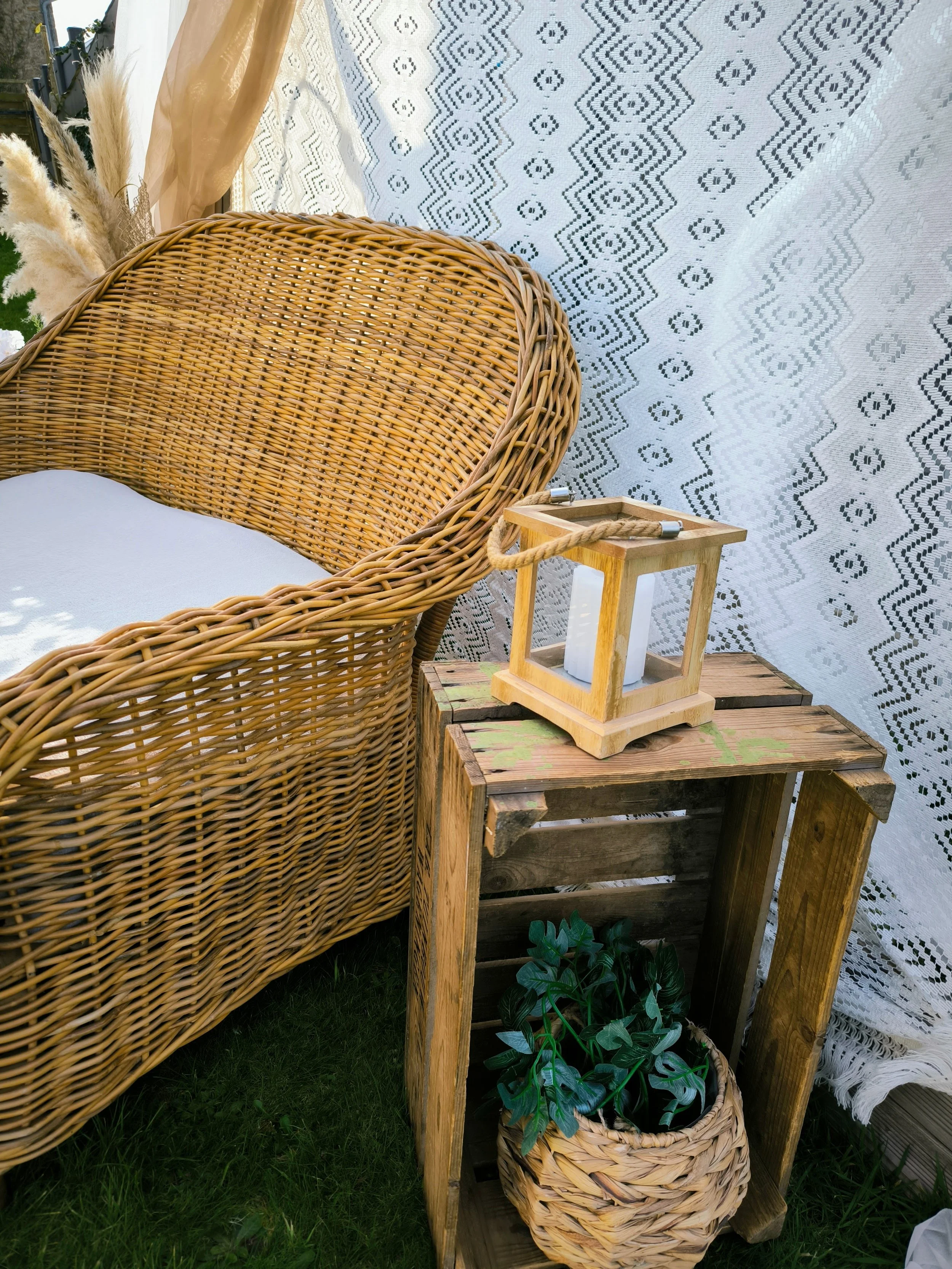 Coin salon avec un fauteuil en rotin, une table en bois rustique avec une bougie dans une lanterne en bois, une plante verte dans un panier en osier. Mur en dentelle en arrière-plan.