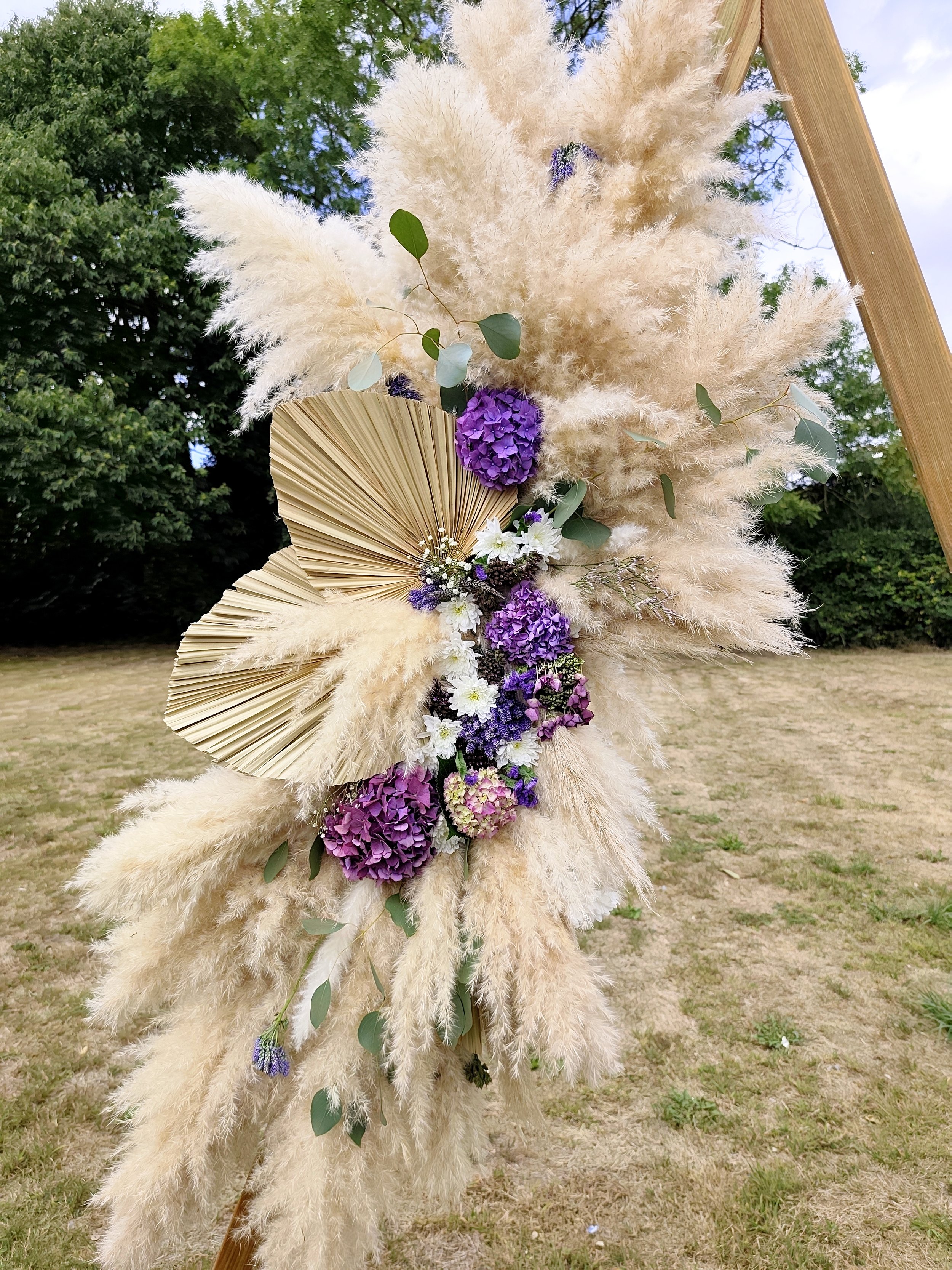 Décoration florale composée de pampas blanches, d'algues vertes, de feuilles de palmier en papier beige, de fleurs violettes, blanches et roses, accrochée à une structure en bois dans un environnement extérieur.