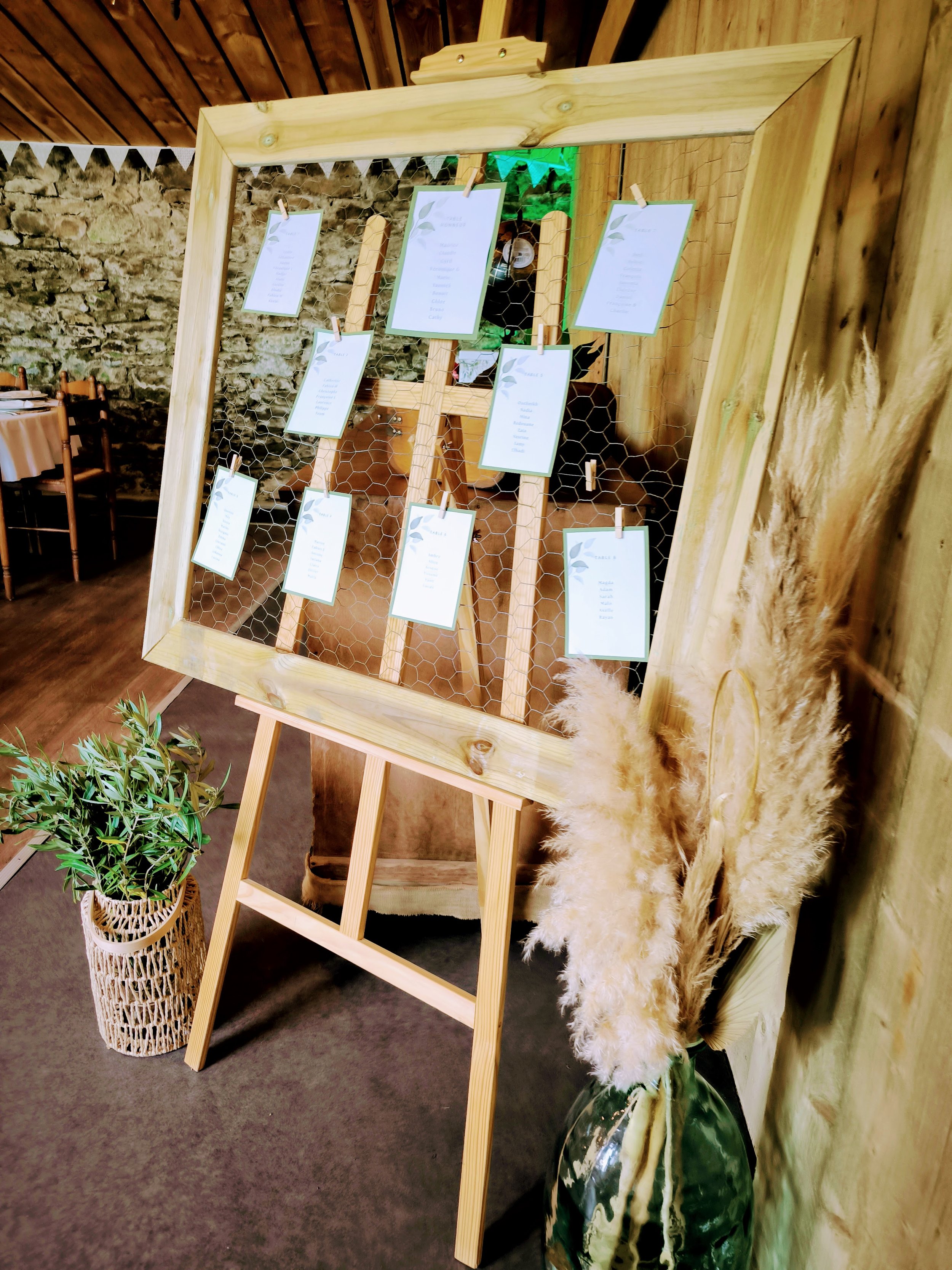 Tableau de plan de table de mariage avec des étiquettes suspendues par des pinces sur un cadre en bois, décorations naturelles avec des pampas et une plante verte dans un vase en céramique.