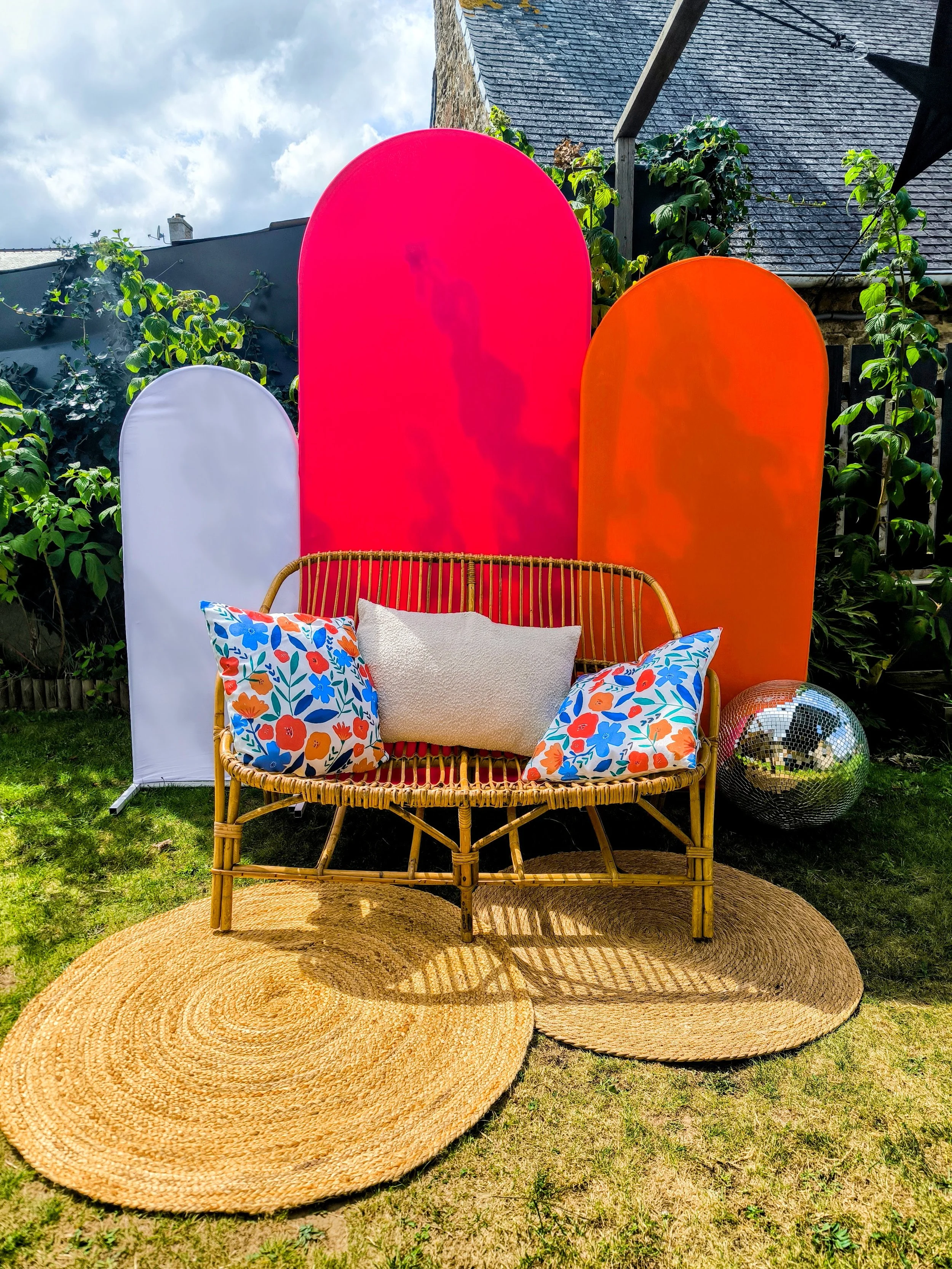 Un coin de jardin avec un canapé en rotin, des coussins colorés et un tapis rond en jute, avec une décoration composée de panneaux en tissu coloré et un miroir disco