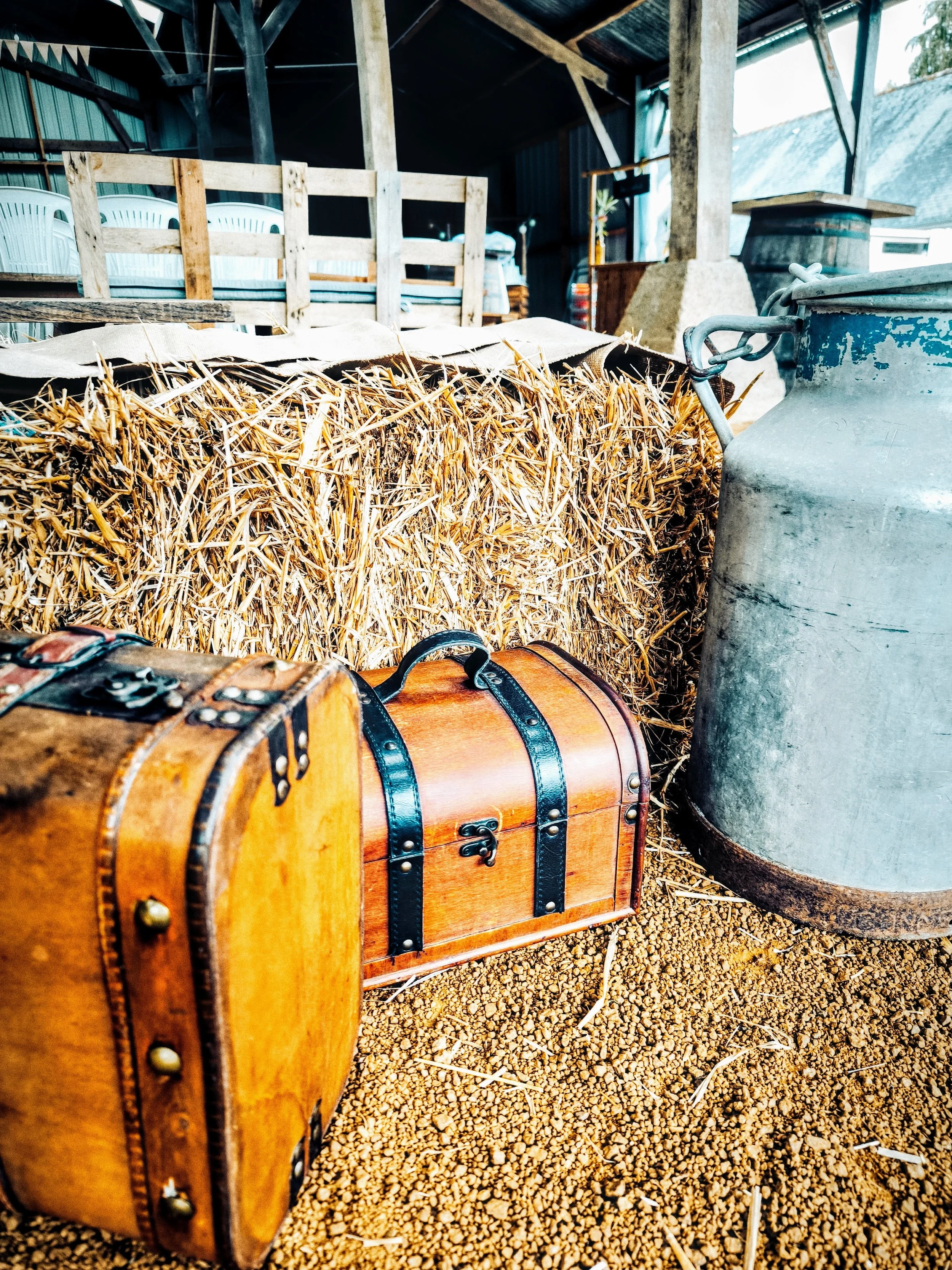 Deux mallettes en bois avec des bretelles en métal, une grande bouse d'étable en métal, et une balle de foin, dans un hangar agricole.