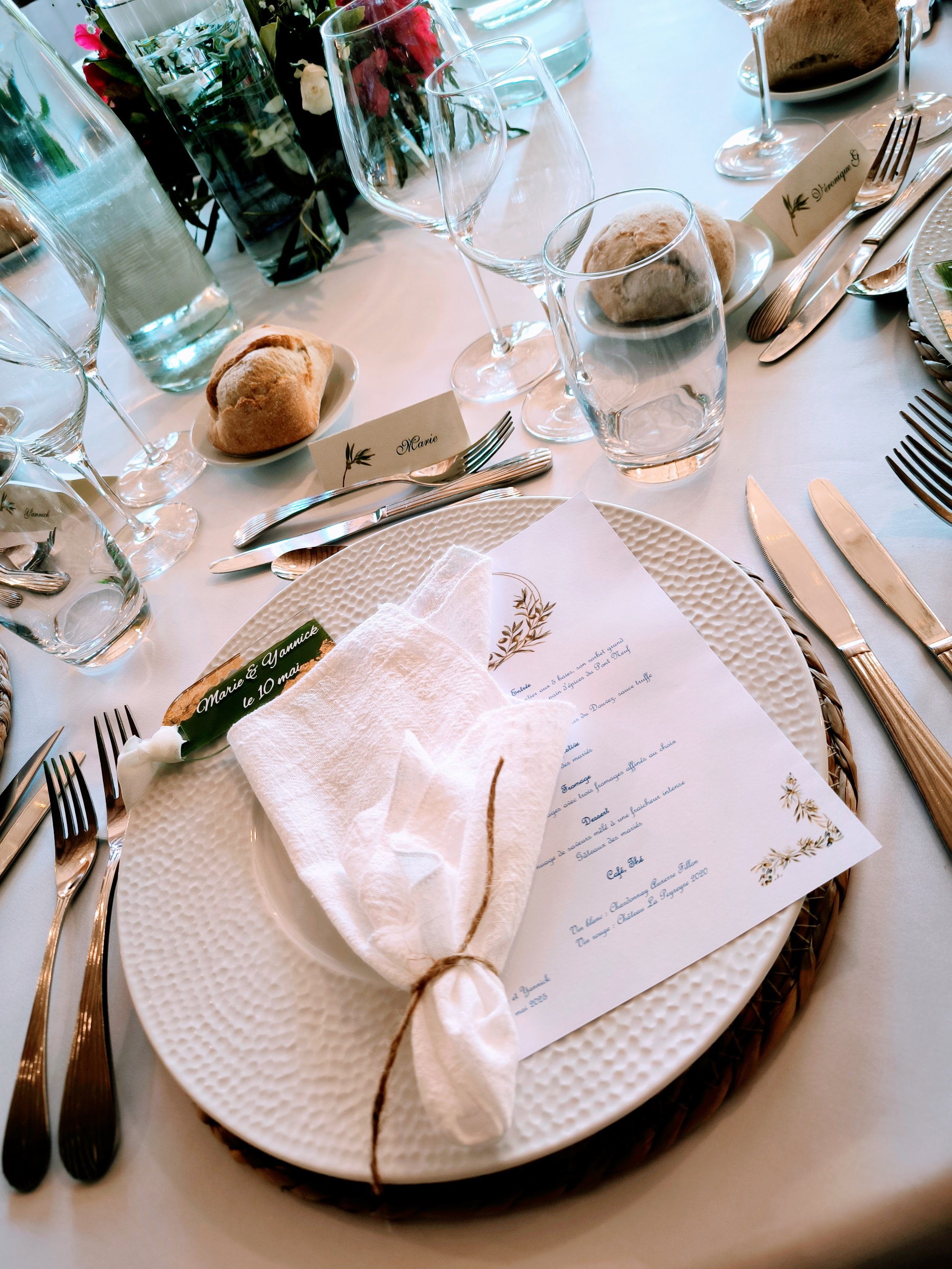 Table dressée pour un repas de mariage, avec une assiette blanche, un menu, une serviette roulée attachée avec une ficelle, et des verres à vin et d'eau.