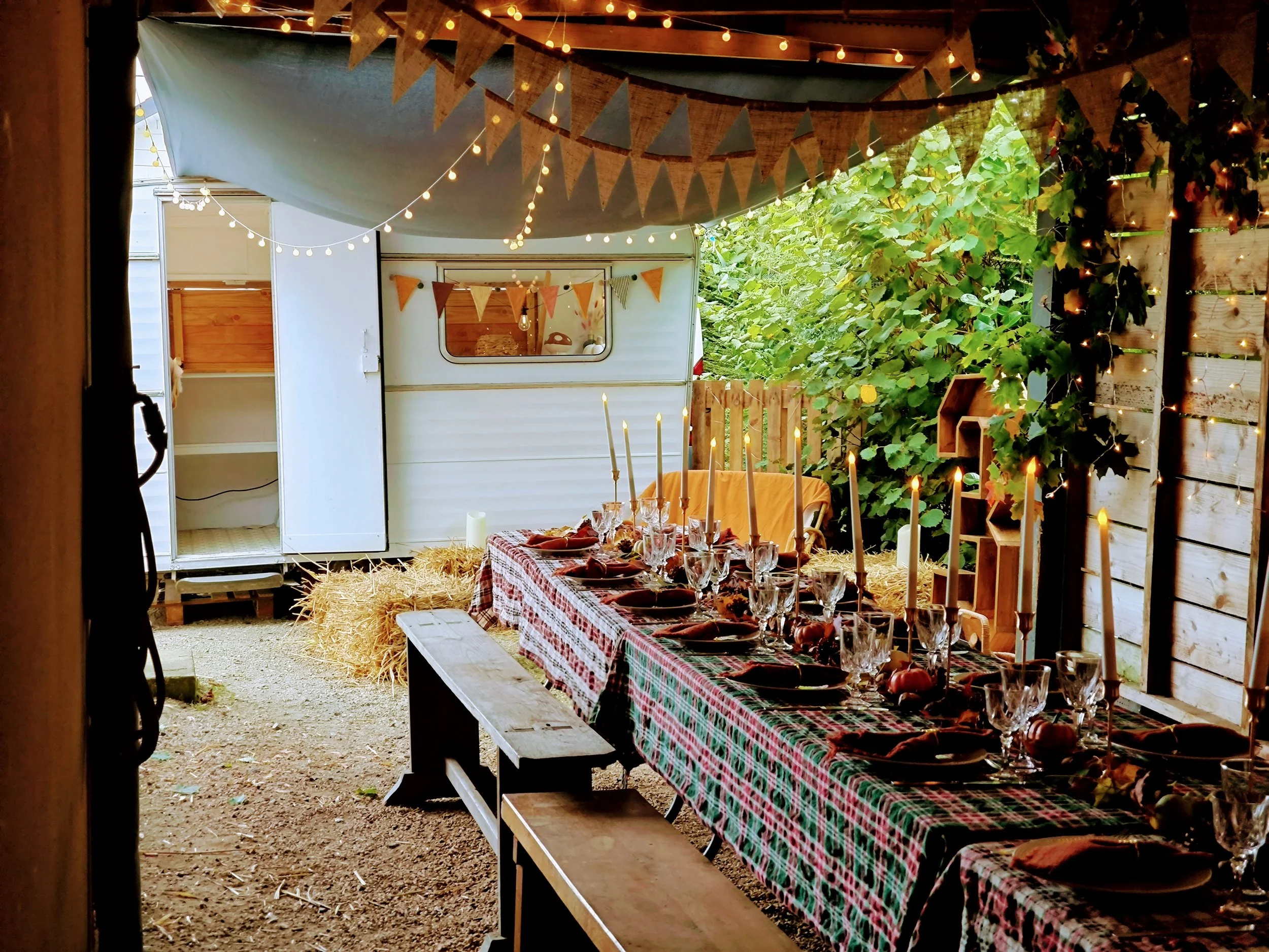 Une table de fête décorée style automnale, avec un chemin de table à carreaux rouges et verts, des bougies, des verres et des assiettes, sous un auvent avec des guirlandes lumineuses et des fanions, caravane photobooth, entourée de verdure.