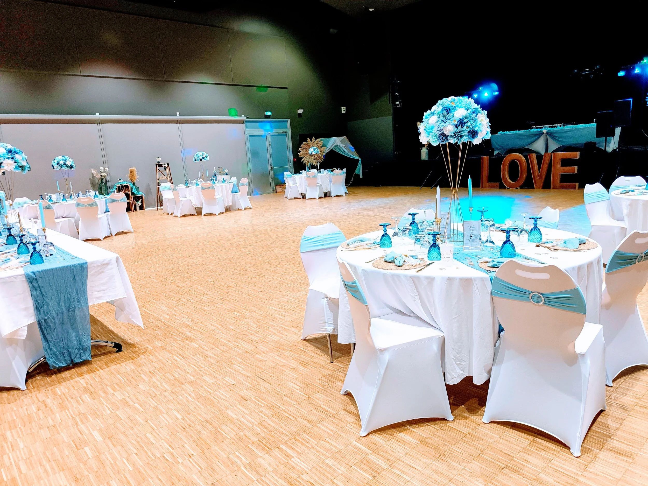 Salle de réception décorée en blanc et bleu avec des tables rondes, des chaises avec des rubans bleus, et des centres de table avec des bouquets de fleurs bleues, avec un grand panneau lumineux formant le mot LOVE à l'arrière-plan.