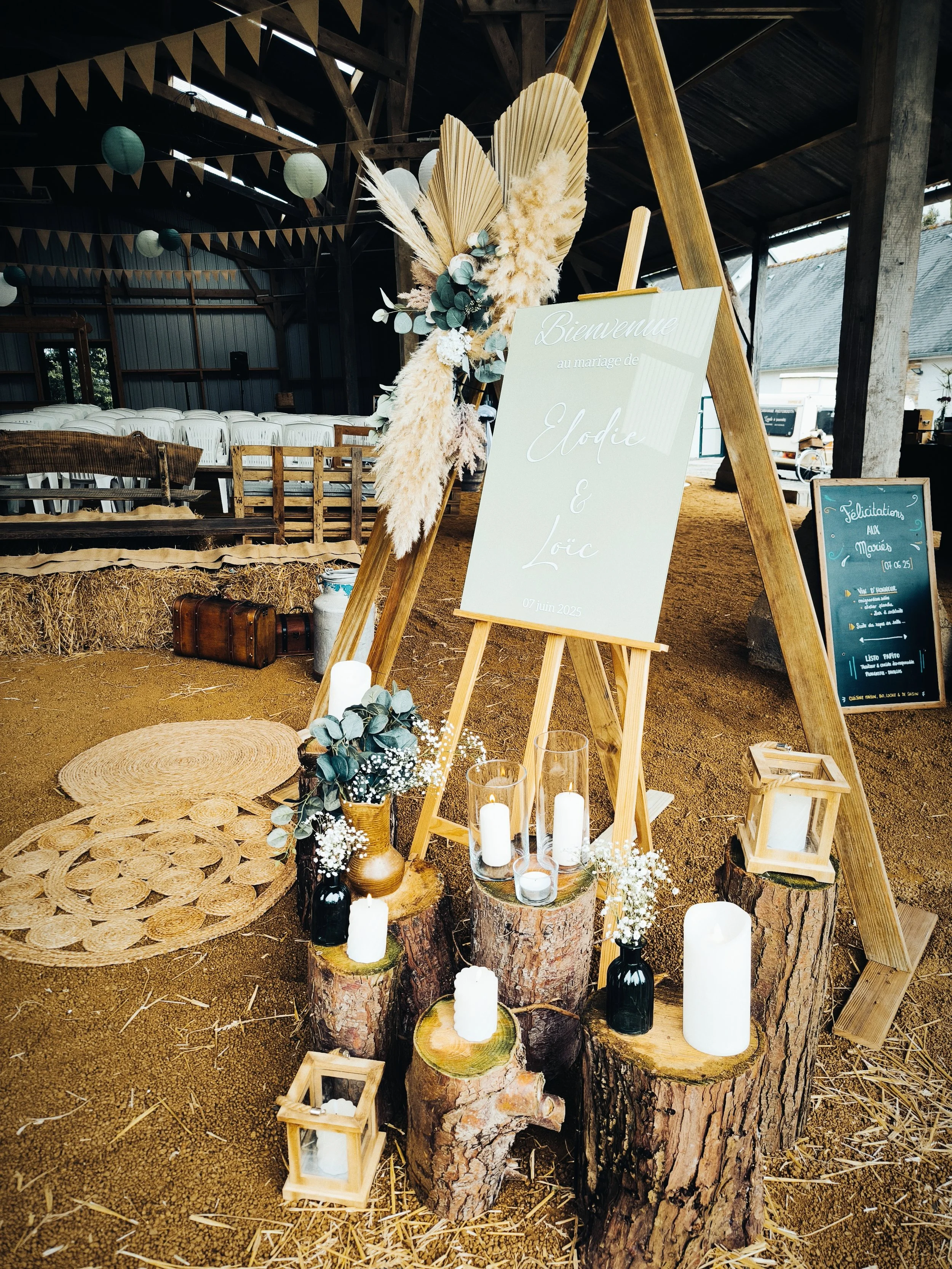 Décoration champêtre pour mariage avec un panneau indiquant les noms des mariés, bougies sur des troncs d'arbres, fleurs séchées et éléments en bois.