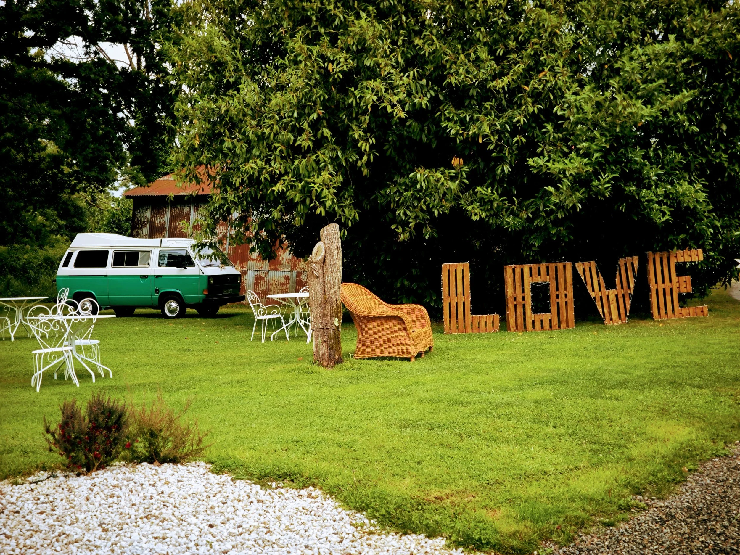 Un jardin avec un véhicule vert, des meubles en fer blanc, une grande enseigne en bois formant le mot 'LOVE', et beaucoup d'arbres et de verdure.
