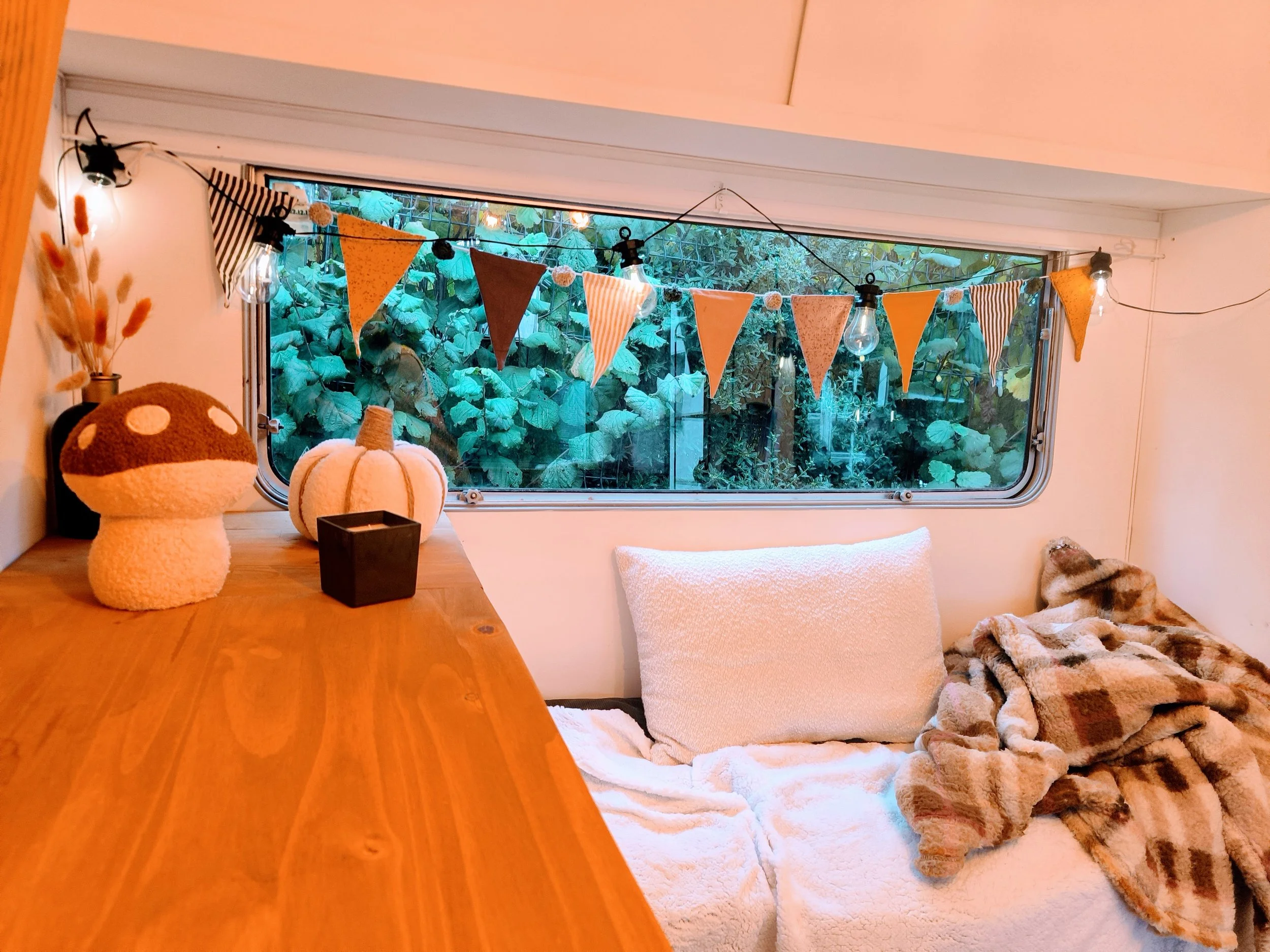Intérieur de la caravane photobooth, une petite pièce confortable avec une fenêtre, décorations de fête en guirlande de triangles, coussins, couverture en fausse fourrure, et décorations automnales sur une surface en bois.