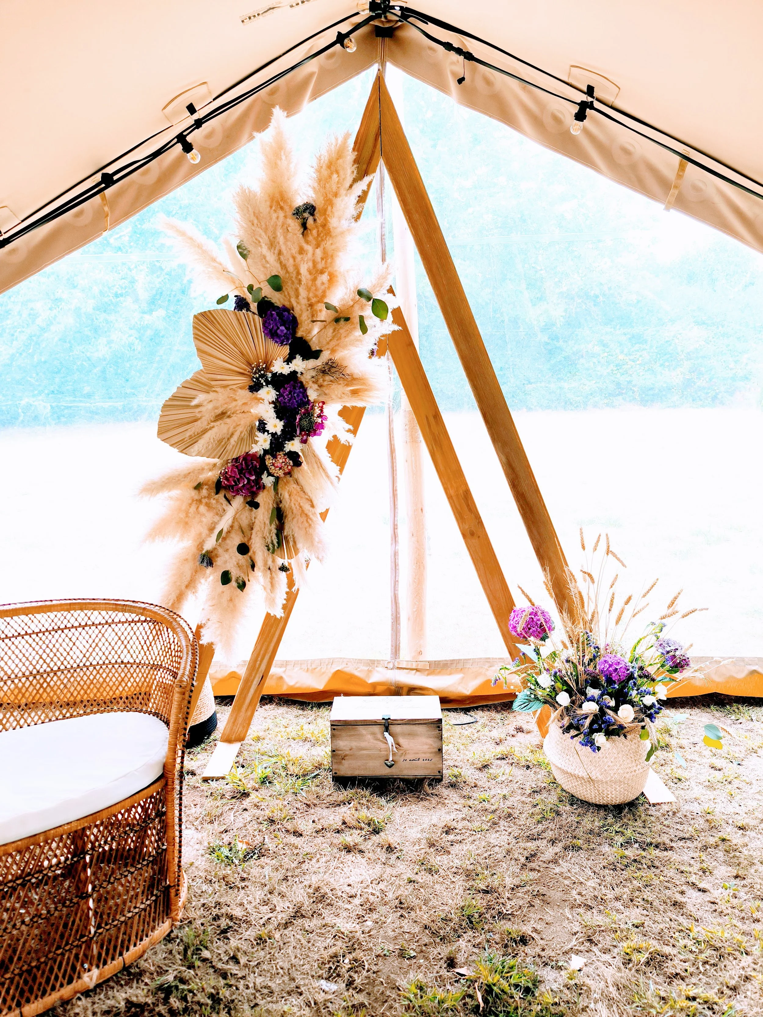 Décoration à l'intérieur d'une tente en toile beige avec un cadre en bois, comprenant une grande composition florale avec des herbes, des plantes séchées, et des fleurs violettes et blanches, un panier de fleurs à l'avant, un coffre en bois au centre
