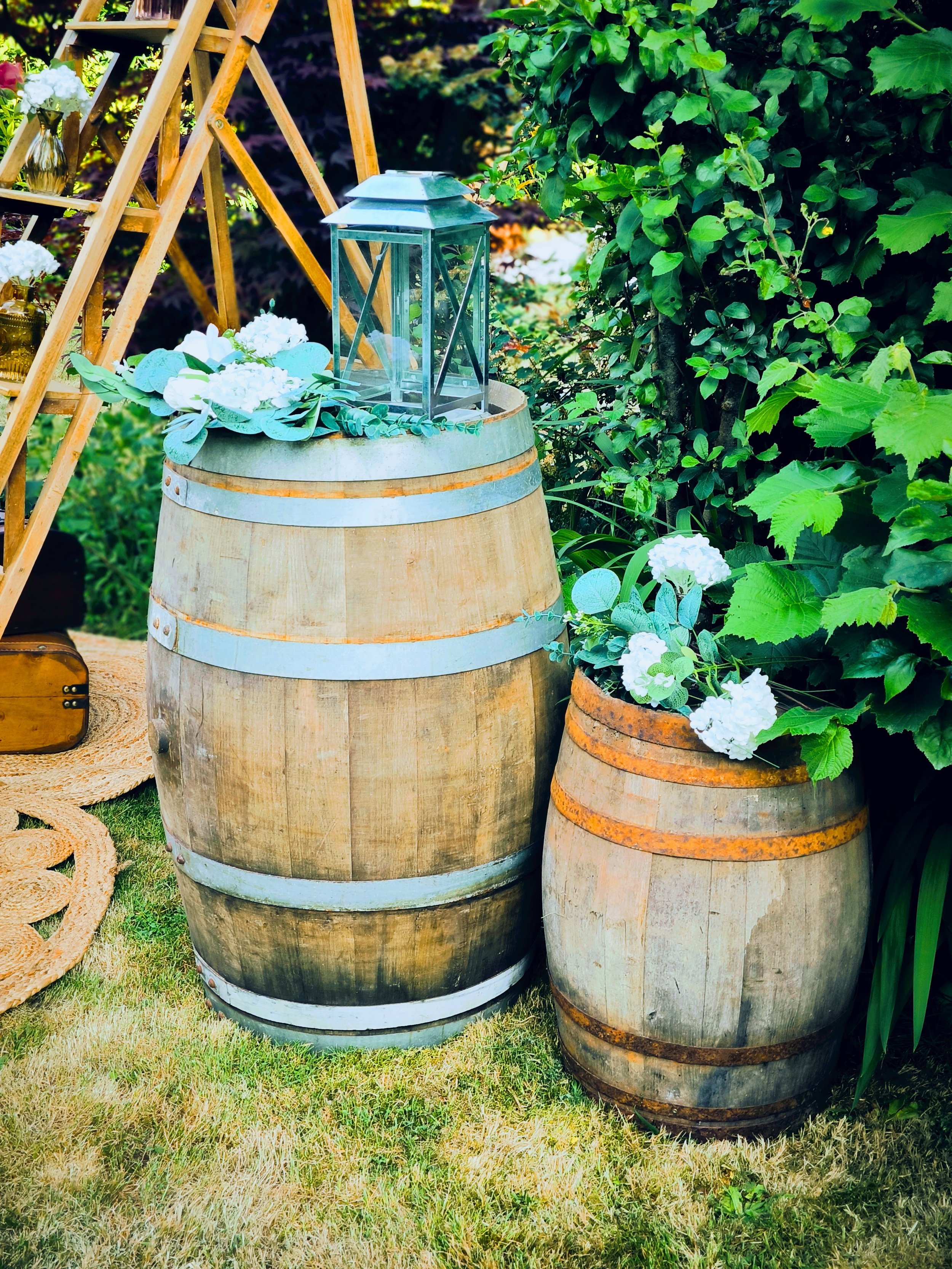 Deux tonneaux en bois transformés en décoration avec des fleurs blanches et un lanterne en verre sur le plus grand tonneau. La scène est entourée de végétation luxuriante.