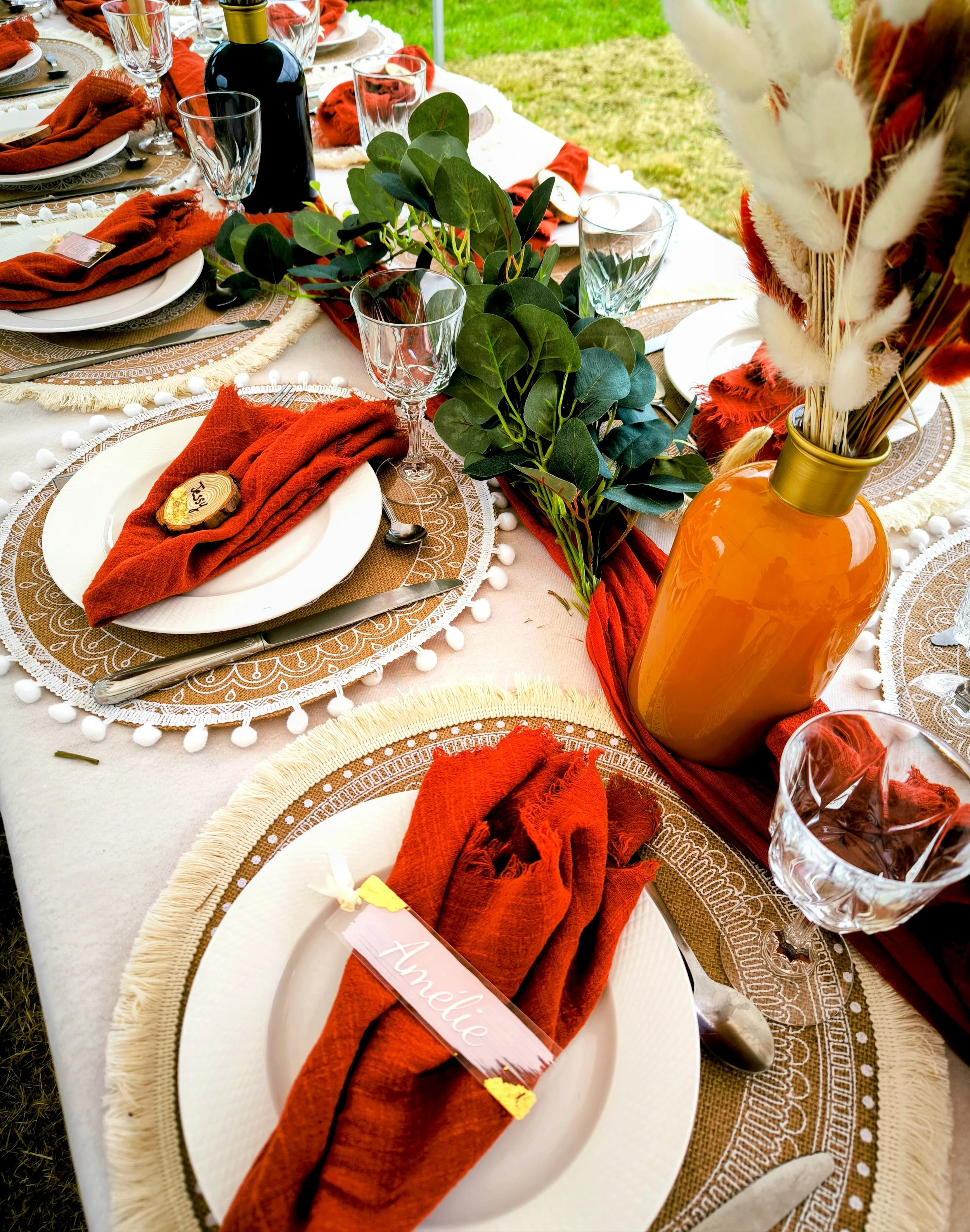 Décoration de table style terra cotta, élégante avec des assiettes, des verres, des couverts, des serviettes rouges/rouille, une branche de feuillage au centre, et un vase orange avec des fleurs colorées.