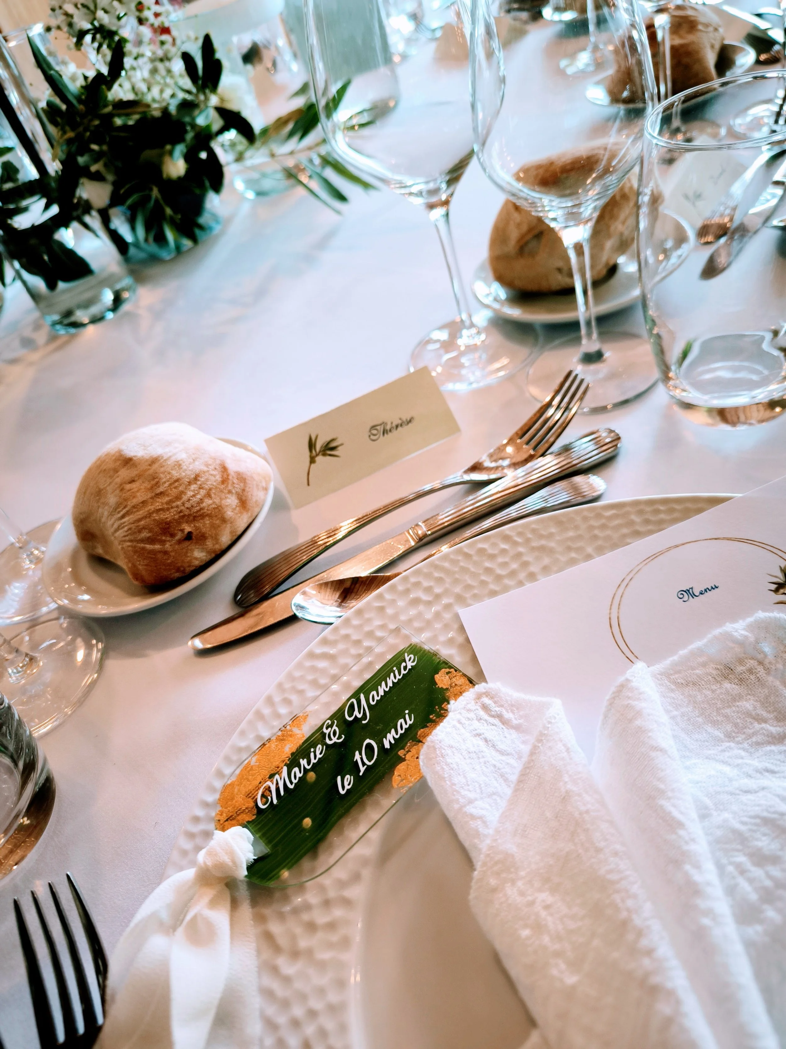 Table de mariage avec des verres, assiettes, assiette avec un rouleau et une petite bouteille de savon ou lotion, menu, et un marque-place indiquant les noms Marie & Yannick et la date 10 mai, décorée avec des fleurs et une nappe blanche.