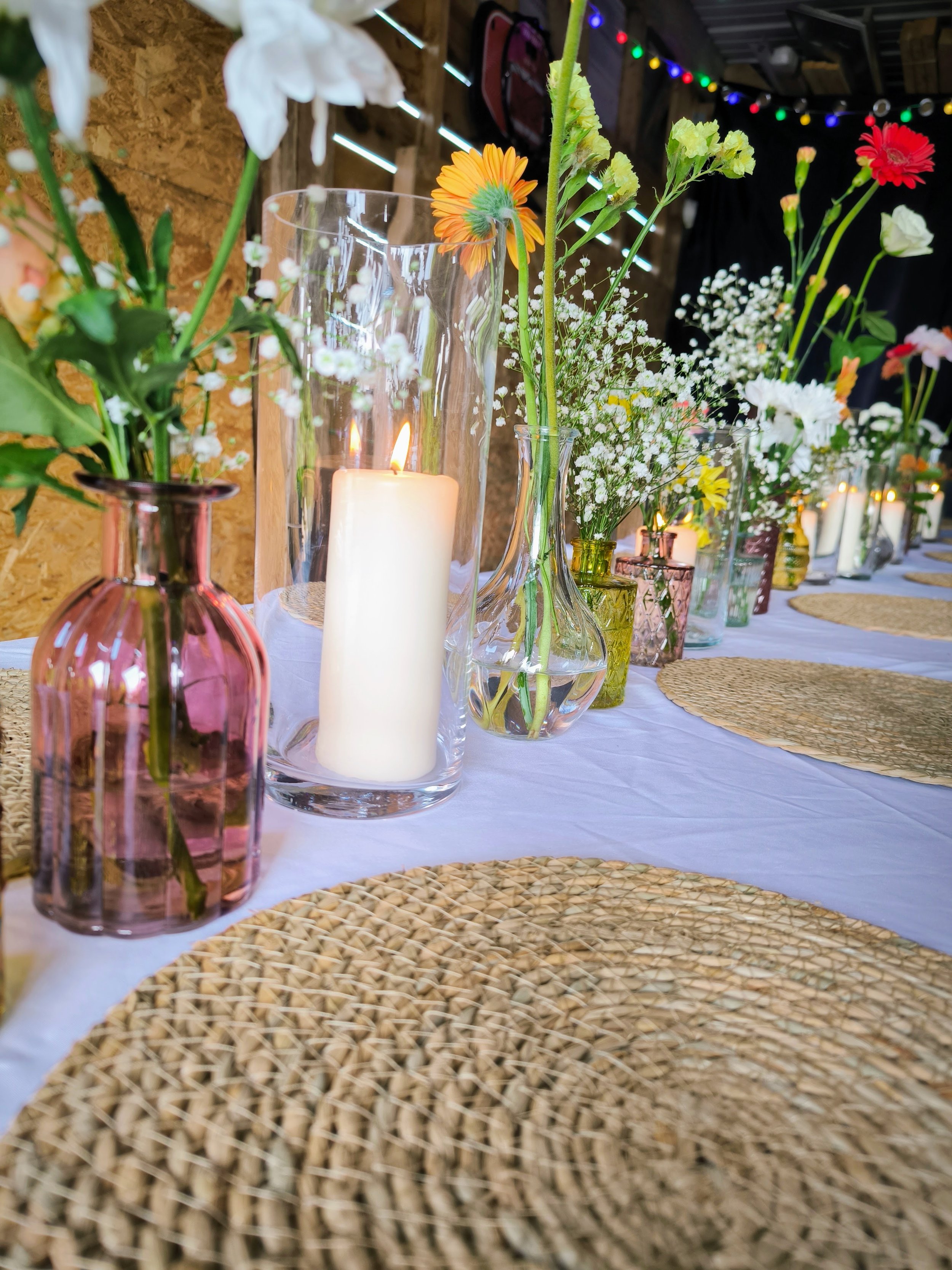 Table décorée avec des vases en verre contenant des fleurs colorées, une bougie blanche dans une grande lanterne en verre, nappes blanches et sets en rotin, ambiance chaleureuse avec des guirlandes lumineuses.