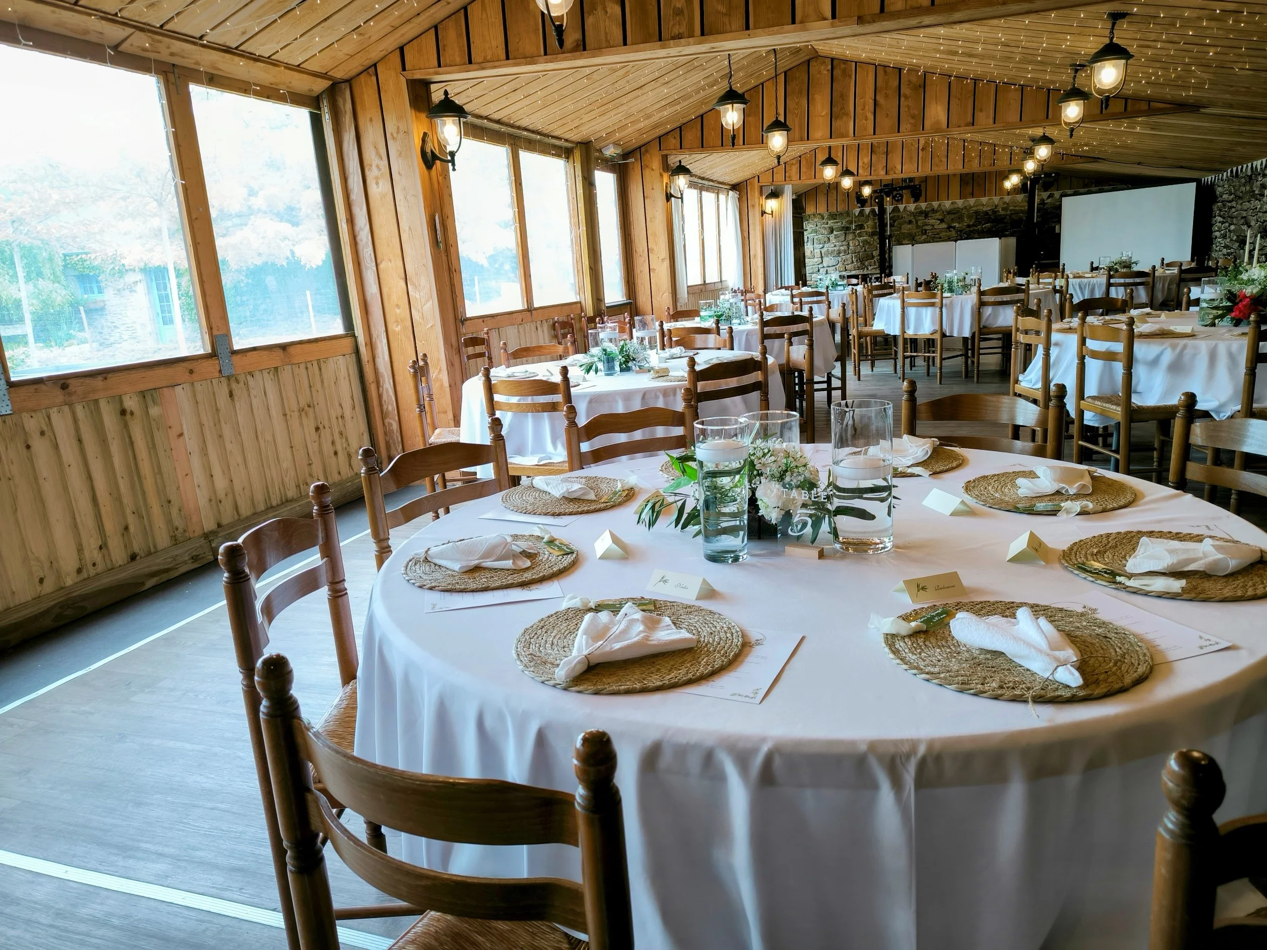 Salle de réception décorée pour un événement avec des tables rondes, nappes blanches, assiettes en osier, verres et centres de table floraux, cheminée en pierre, lumière tamisée et grand écran au fond.