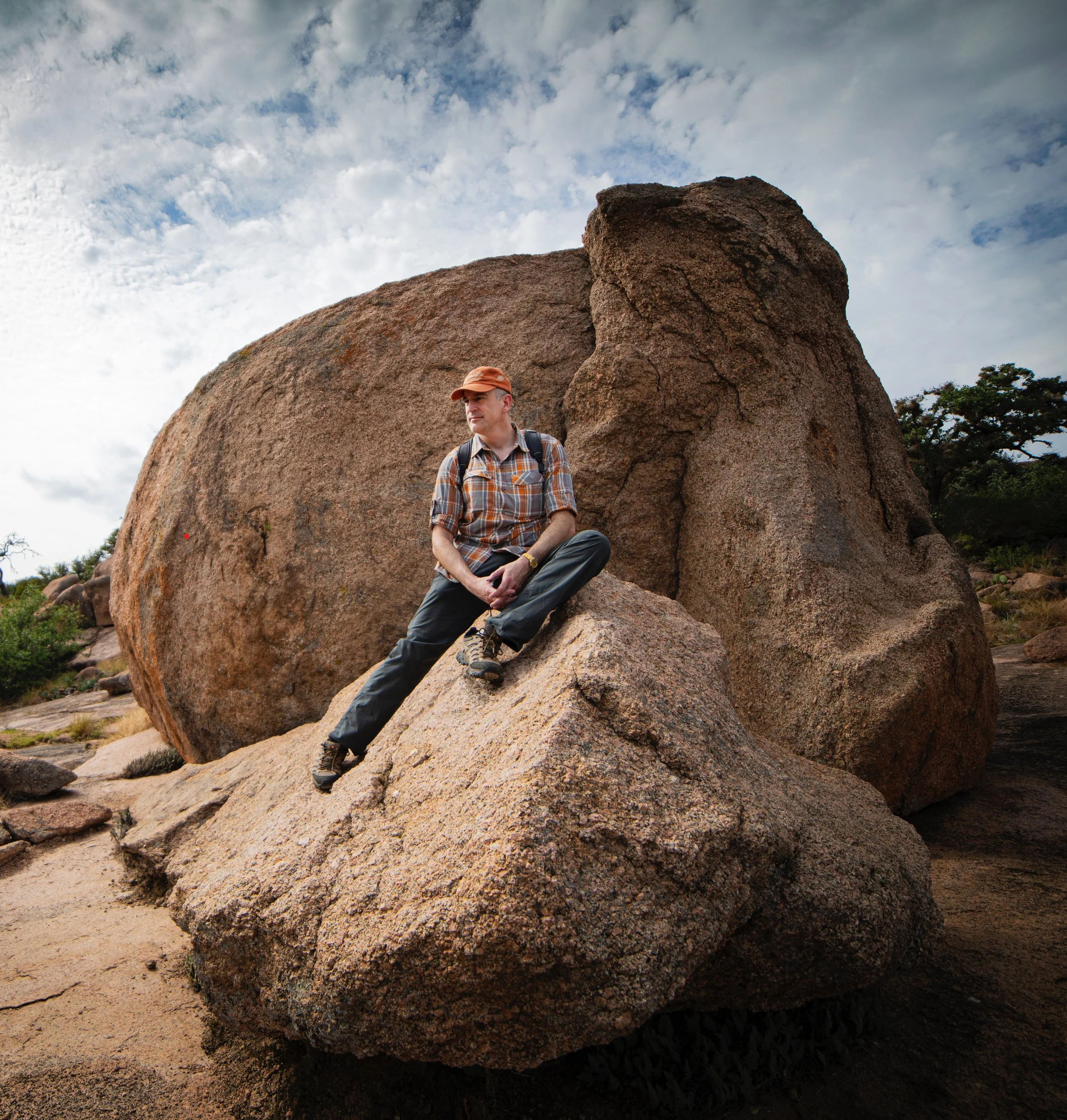 A man sitting on a large rock in a desert landscape with cloudy sky and trees in the background.