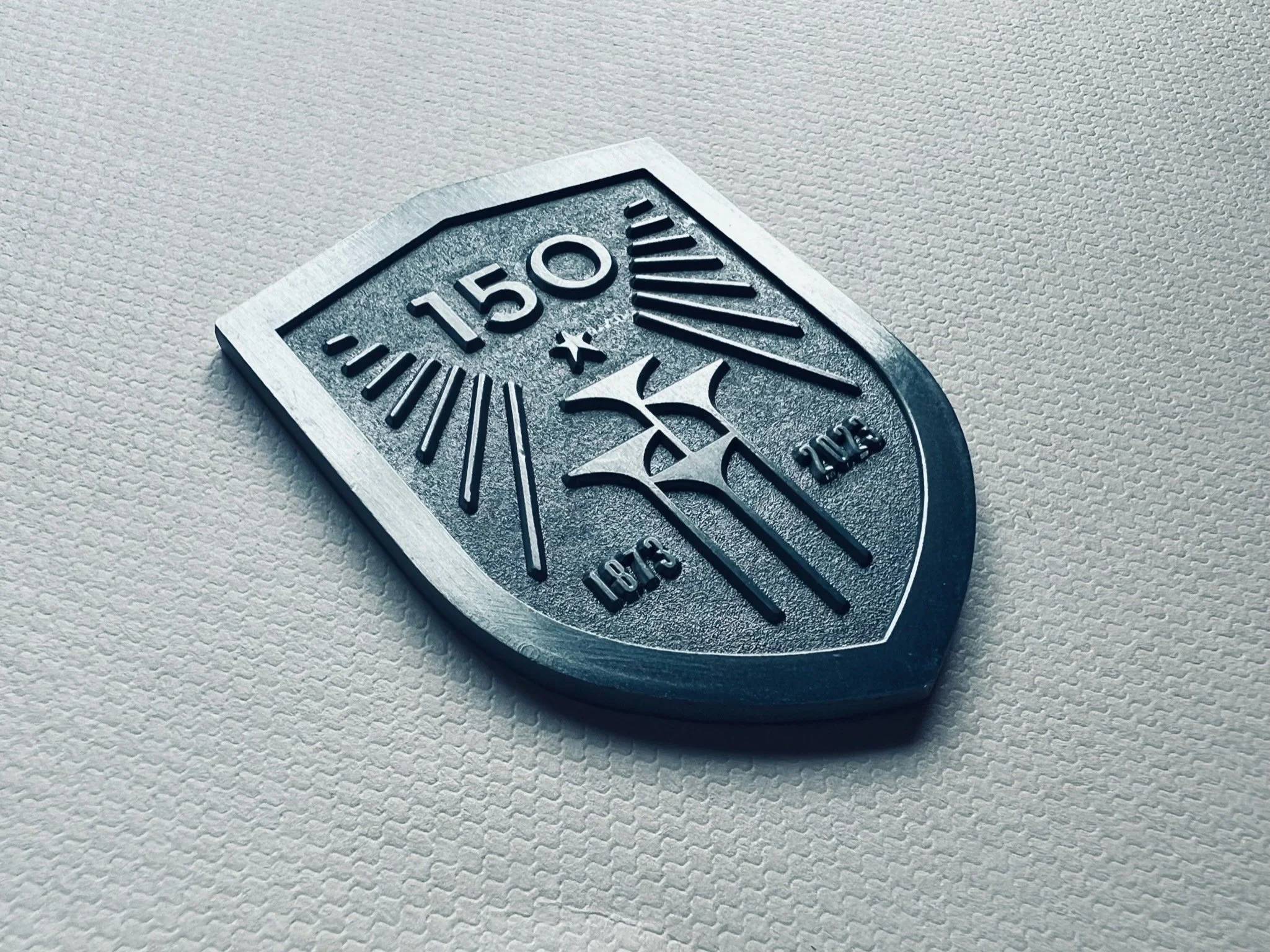 A metallic shield-shaped badge with the Mercedes-Benz logo, inscribed with '150', 'x 2013', '188.16', and '2013', resting on a textured light-colored surface.