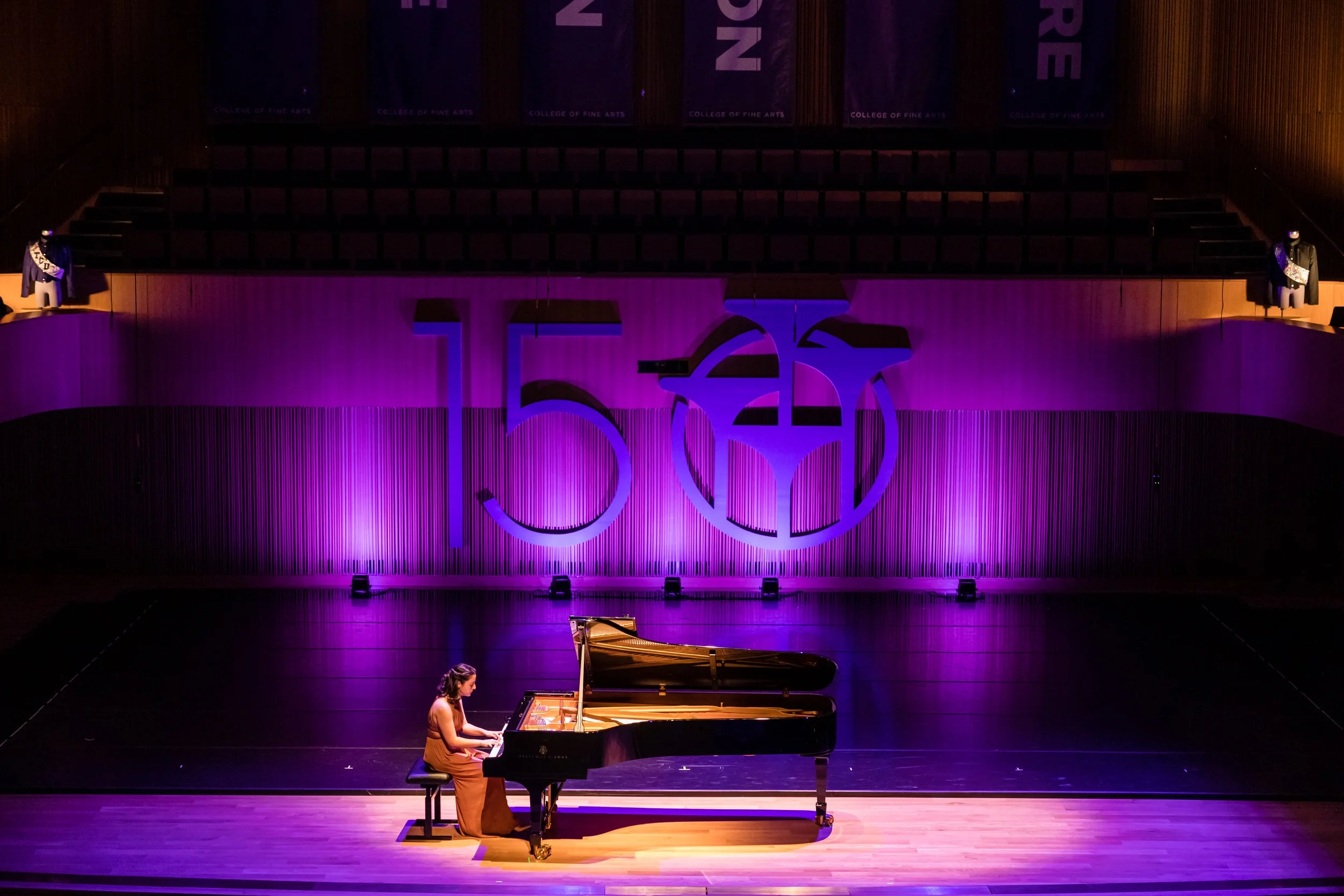 A woman playing a grand piano on stage at a concert hall with purple lighting, and a large '50' number as part of a decoration or backdrop.