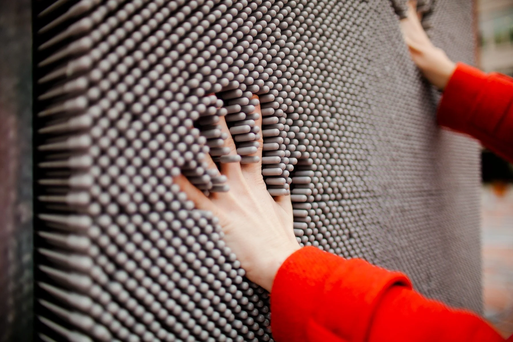 Close-up of hands touching a metal surface with numerous pins or nails, wearing red sleeves.