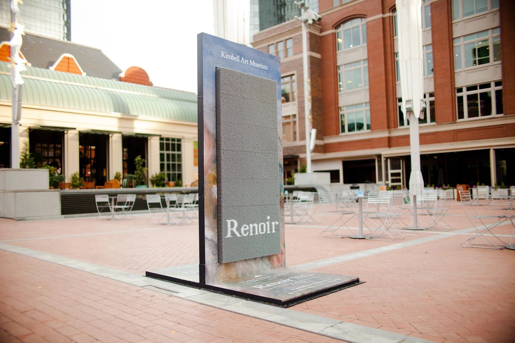 Empty outdoor plaza with metal tables and chairs, a sign for Kimbell Art Museum, and a building in the background with large windows and brick facade.