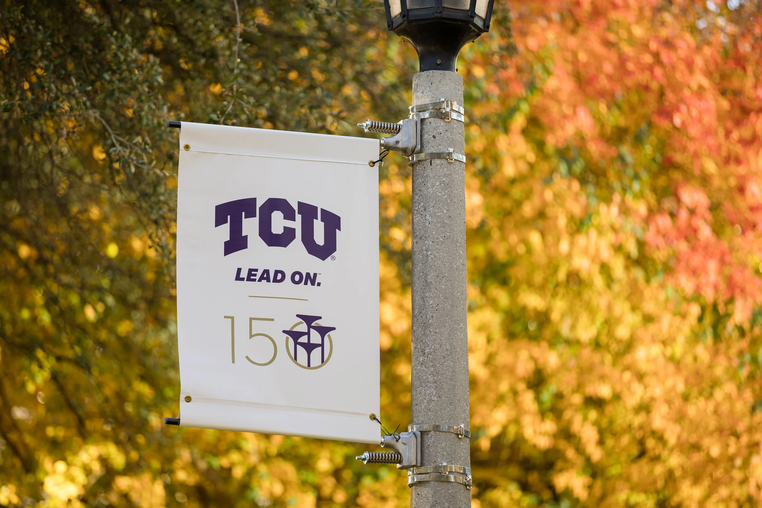 Flag with TCU logo and text urging to lead on 15 near colorful autumn trees.