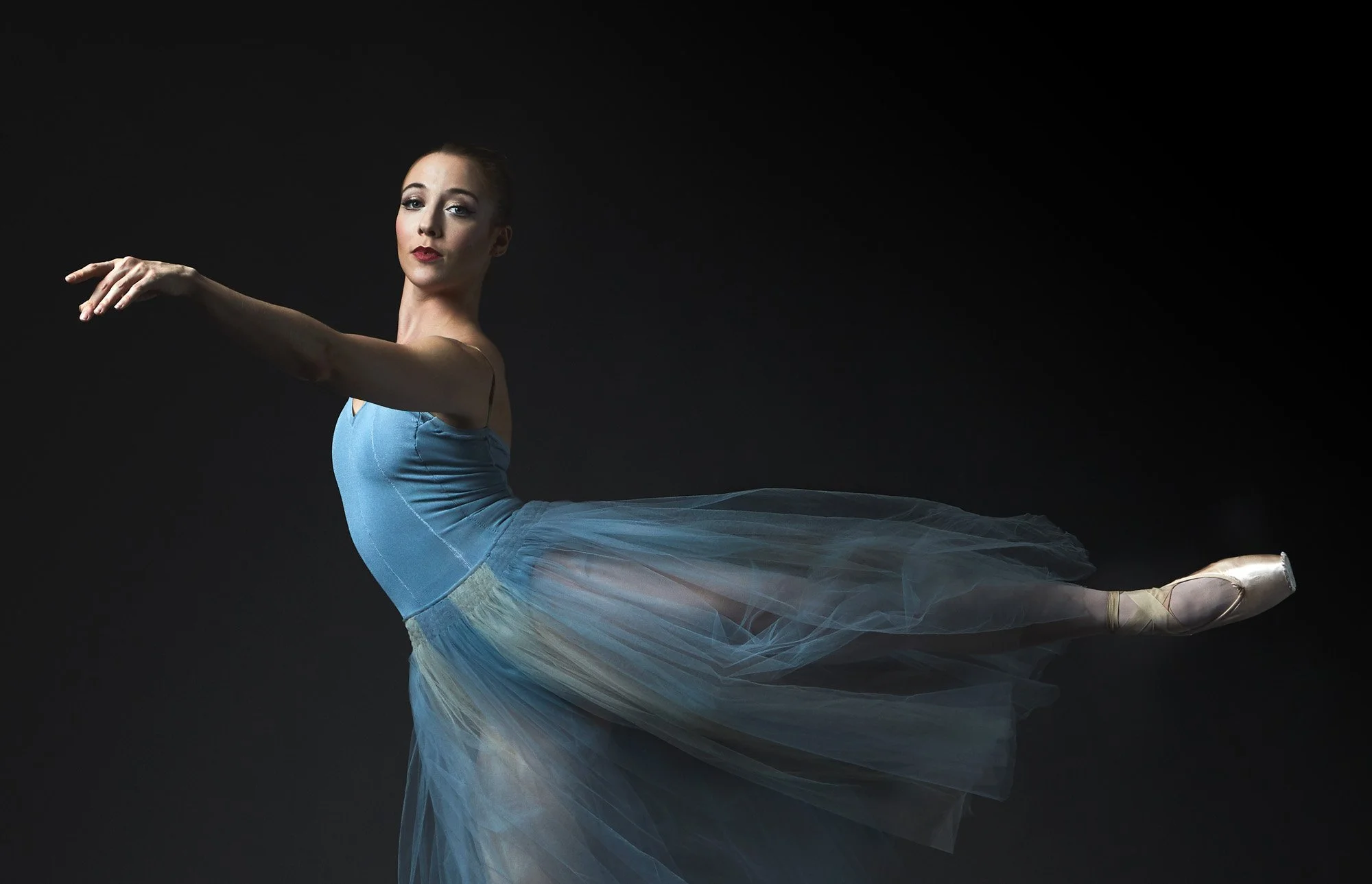 Ballet dancer in a blue leotard and tutu performing a ballet pose against a dark background.