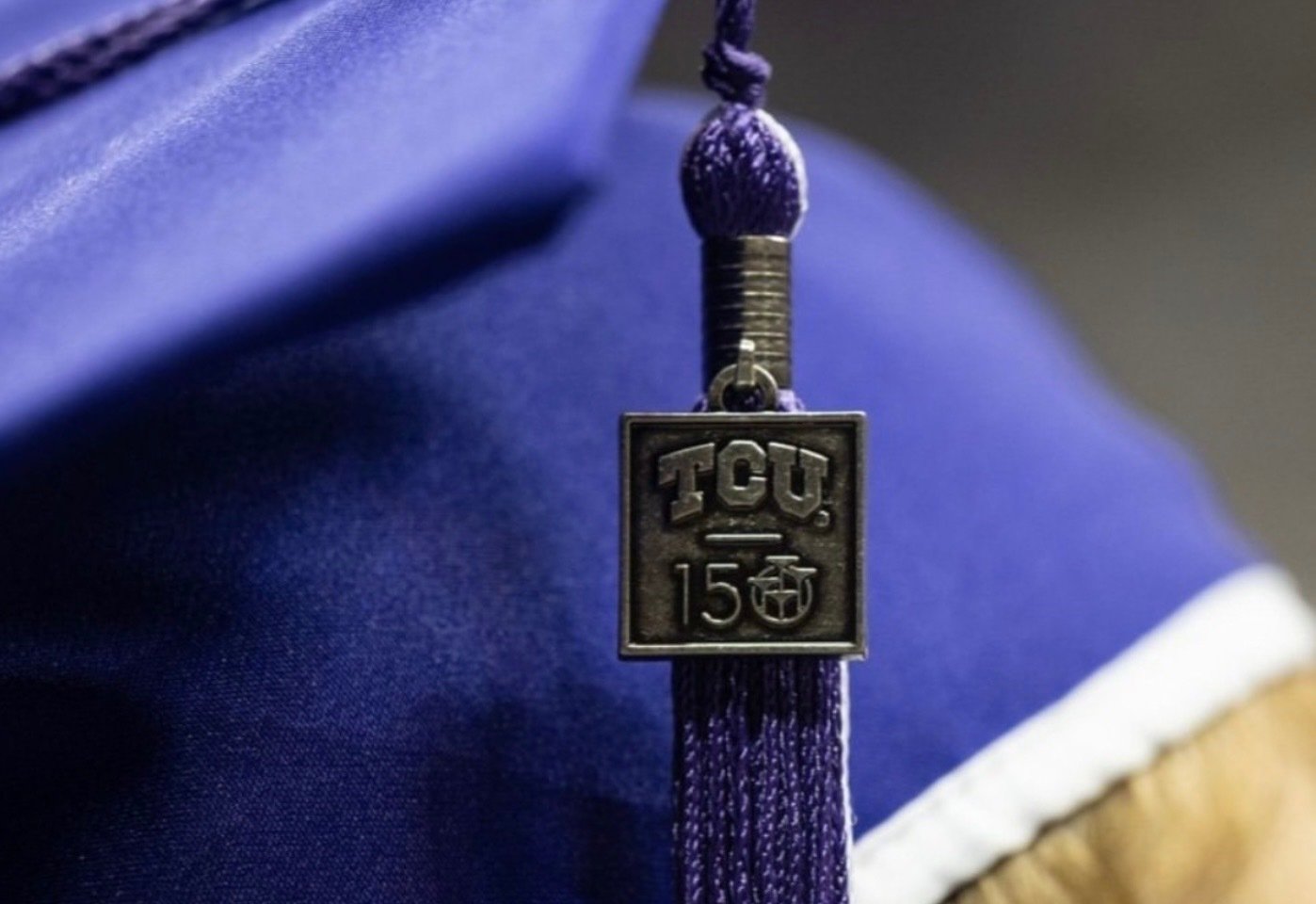 Close-up of a graduation cap's tassel with a metallic charm engraved with 'TCU 150' hanging from a purple cord.