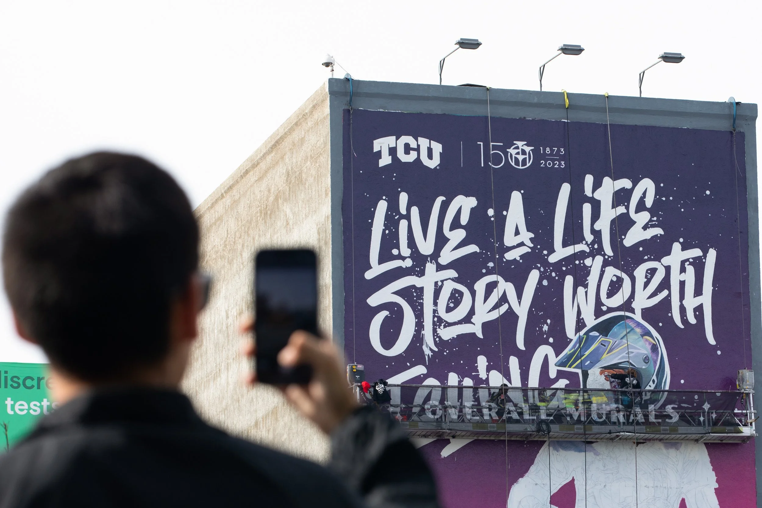 A person taking a photo of a large mural. The mural has the text "LIVE A LIFE STORY WORTH LIVING" and features the TCU logo and a graphic of a train. The mural commemorates TCU's 150th anniversary from 1873 to 2023.