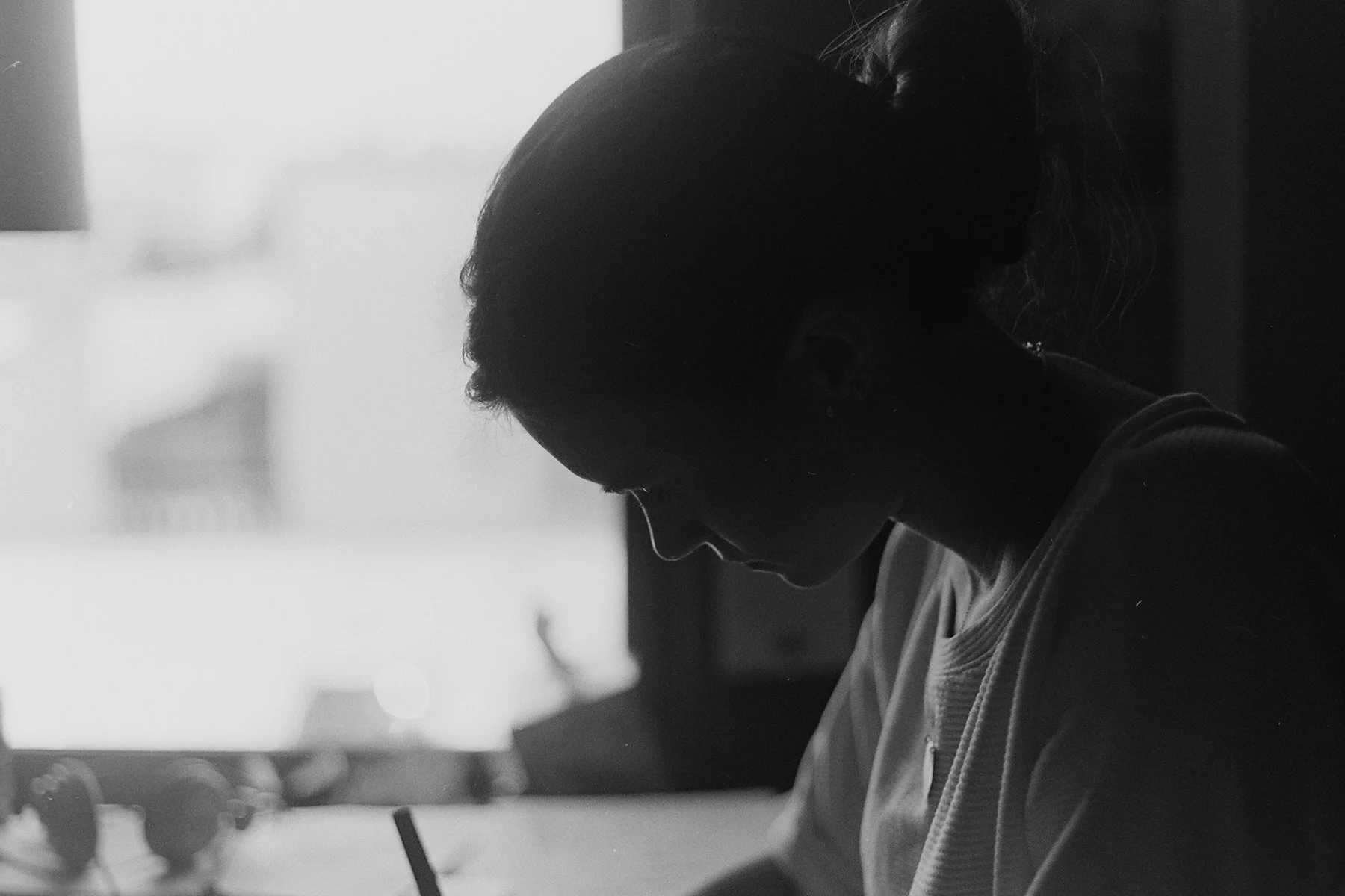 Silhouette of a woman with a ponytail, looking down and writing, with a window in the background.