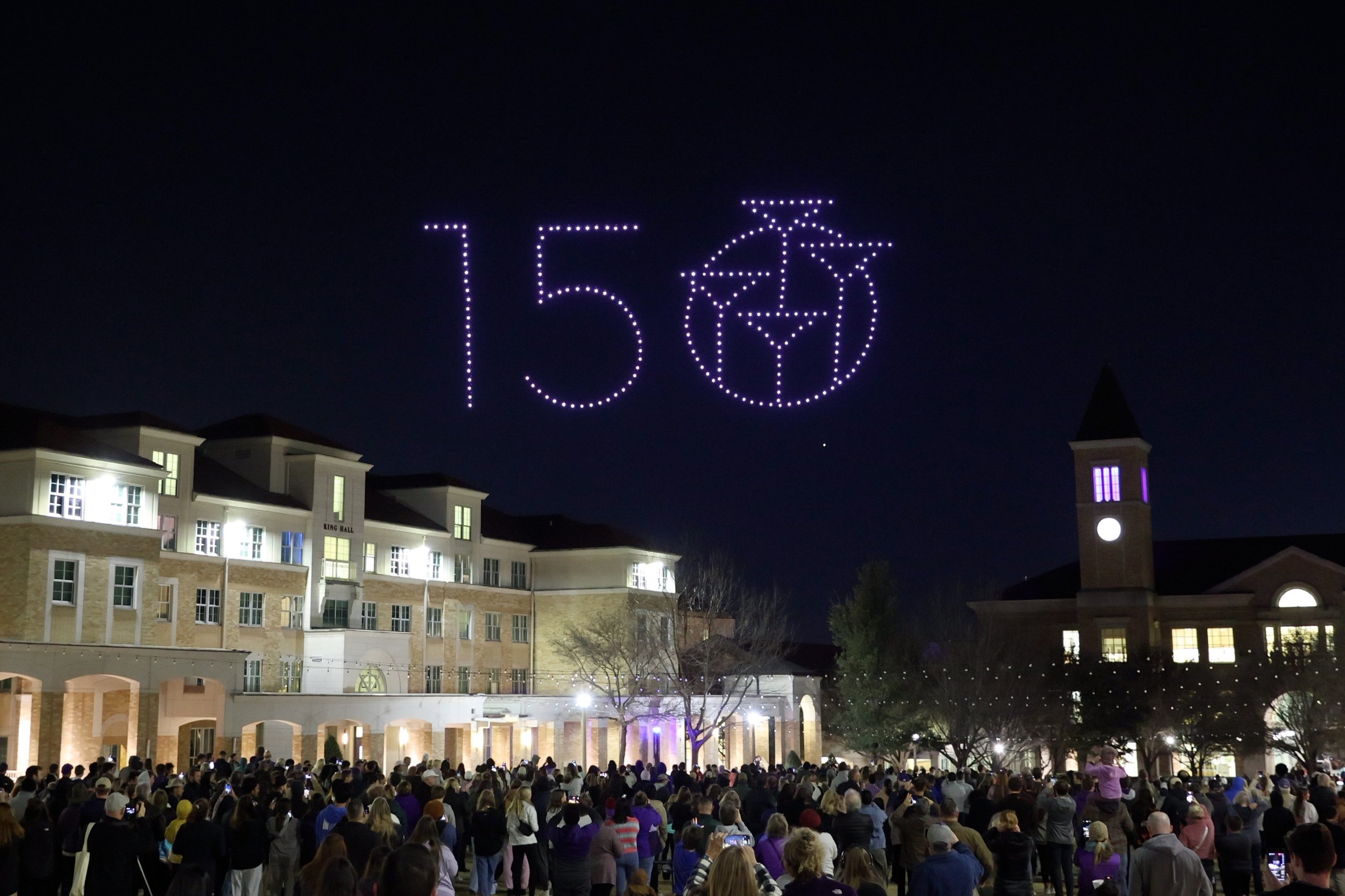Large crowd gathered outside in front of a building at night, with a sky display of the number 150 and a nautical-themed circle with a sailboat and map elements illuminated in purple lights.